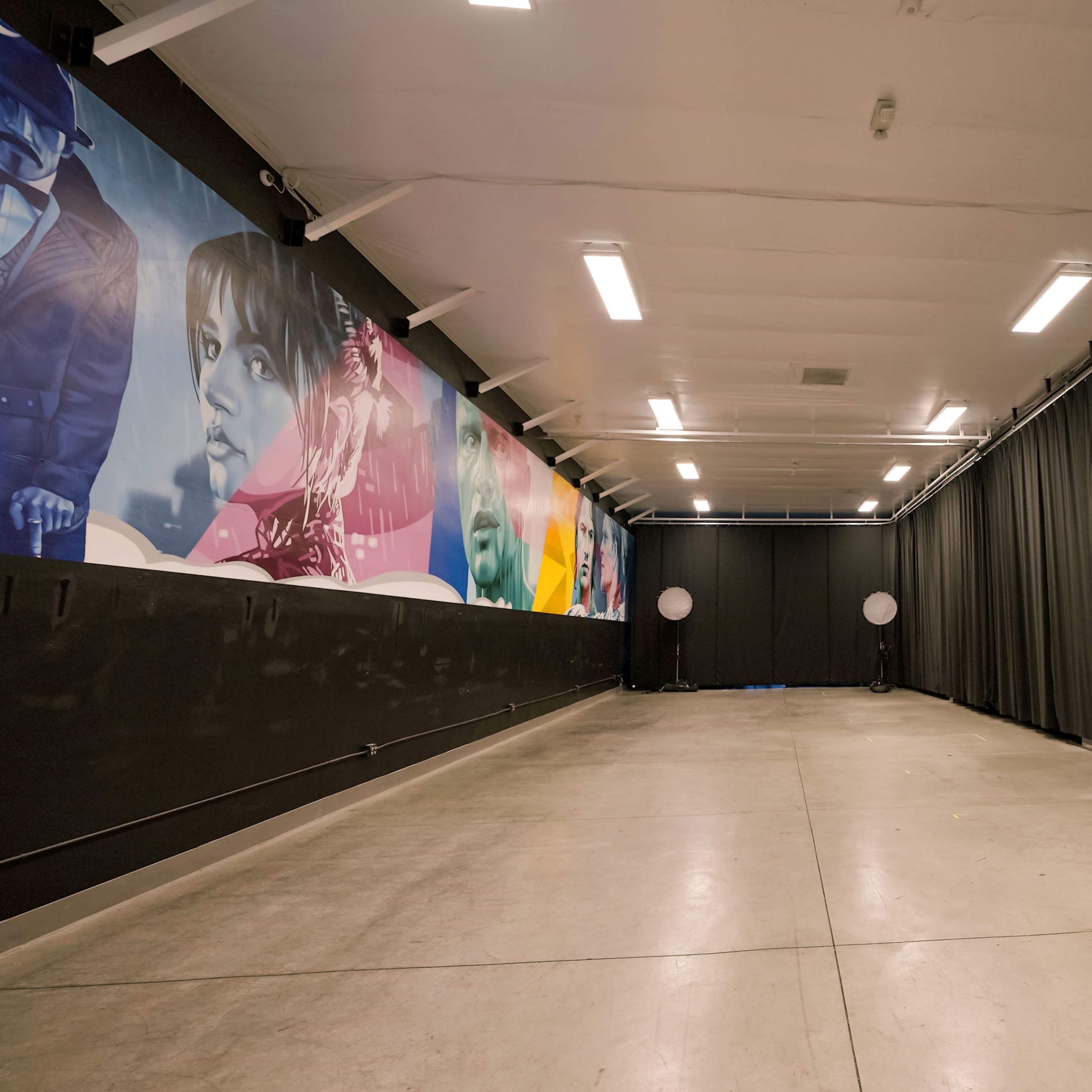A spacious indoor studio with a concrete floor, black curtains lining the walls, and a colorful mural on one side.