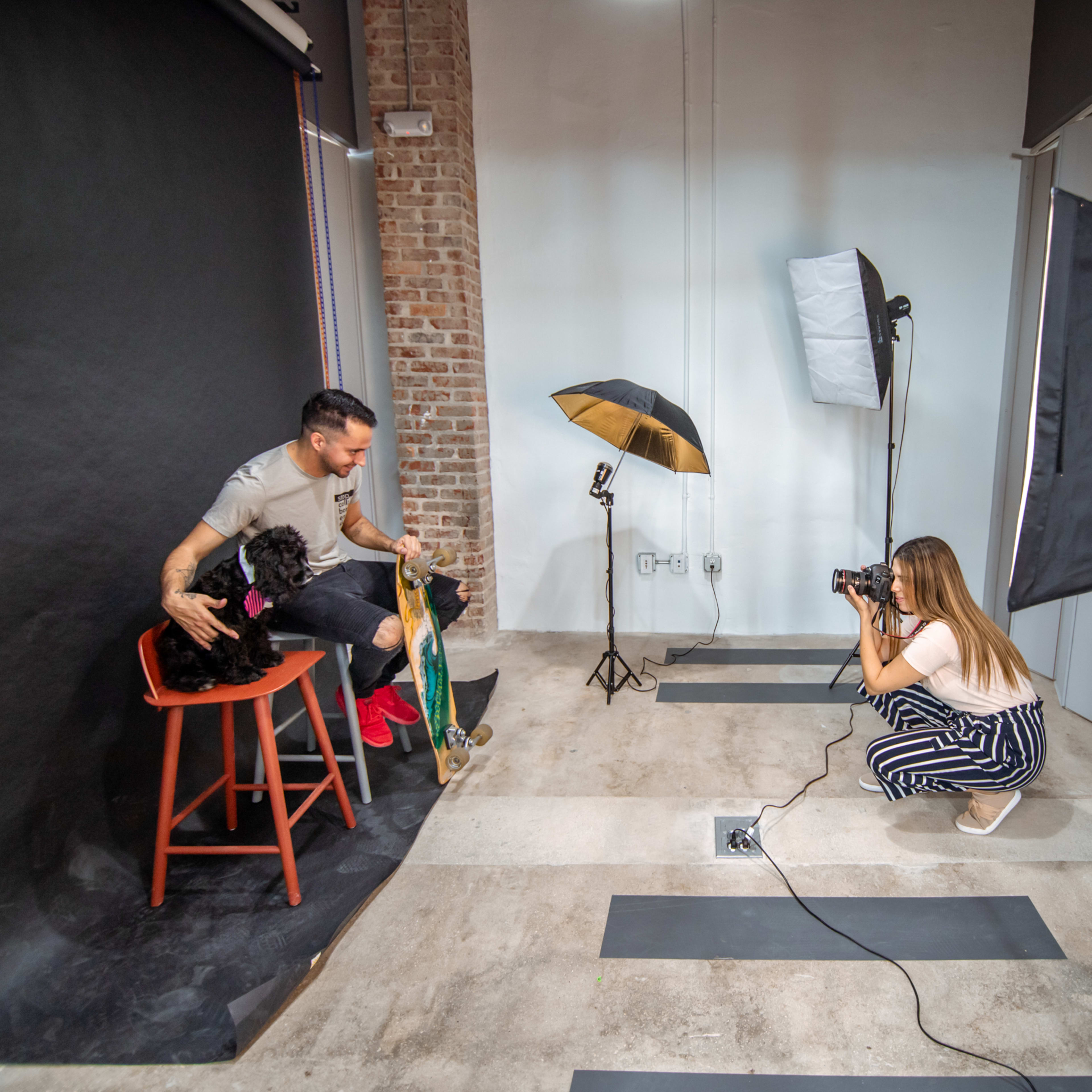 A photographer captures images of a man holding a dog while sitting on a stool in a studio setup with lighting equipment.