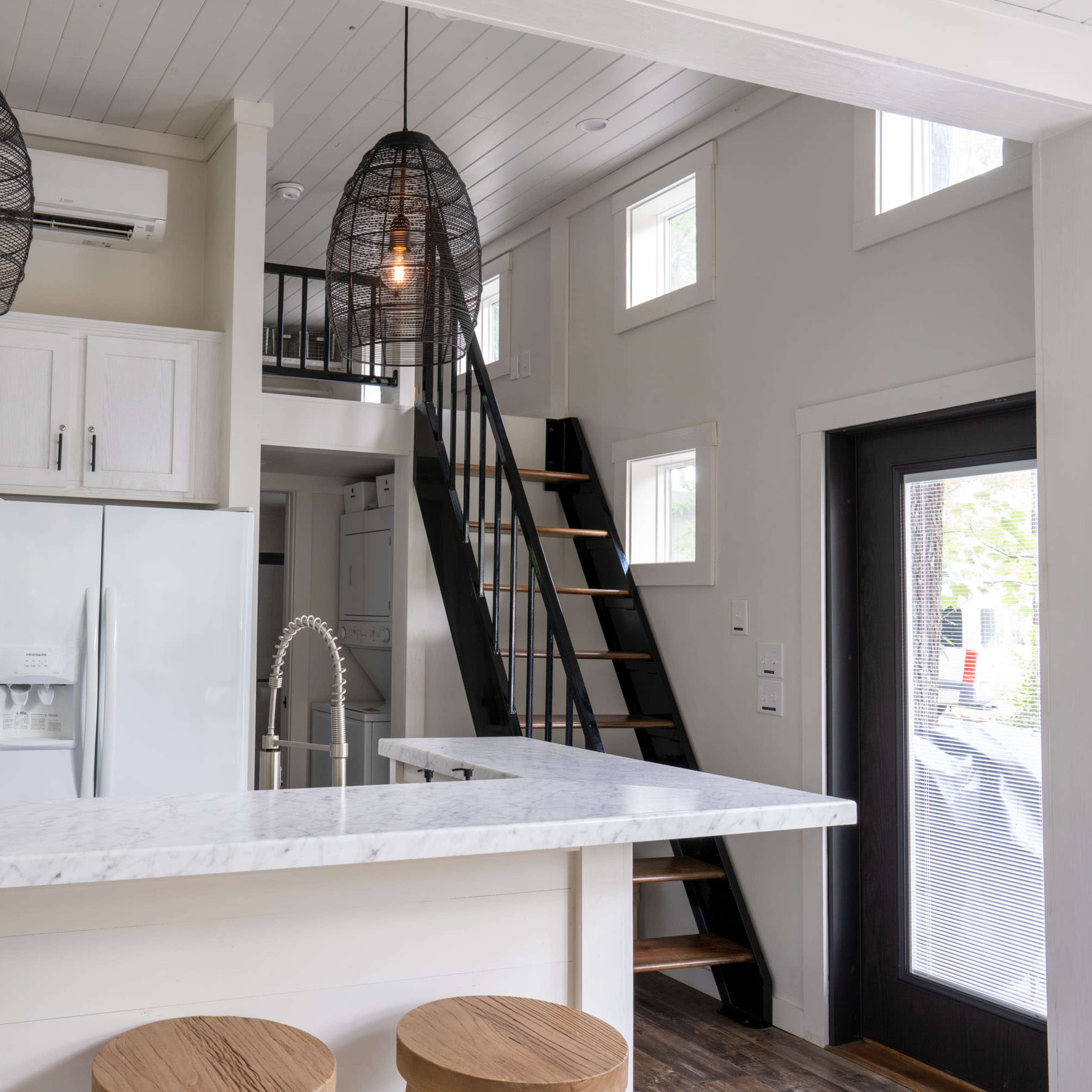 A modern kitchen area with a marble countertop, wooden bar stools, and a staircase leading to an upper level.