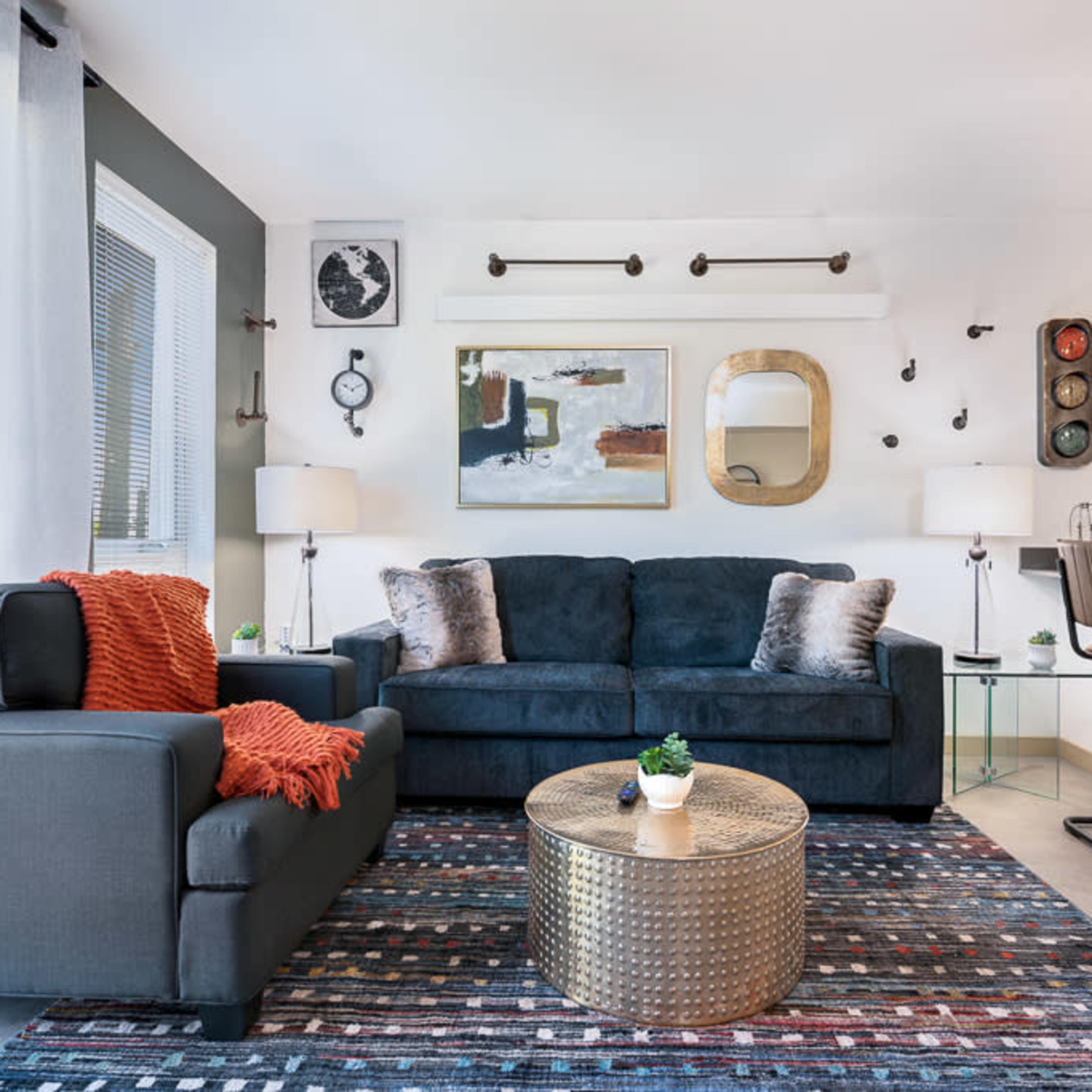 A modern living room with a dark sofa and armchair, a round metallic coffee table, artistic wall decor, and large windows allowing natural light.