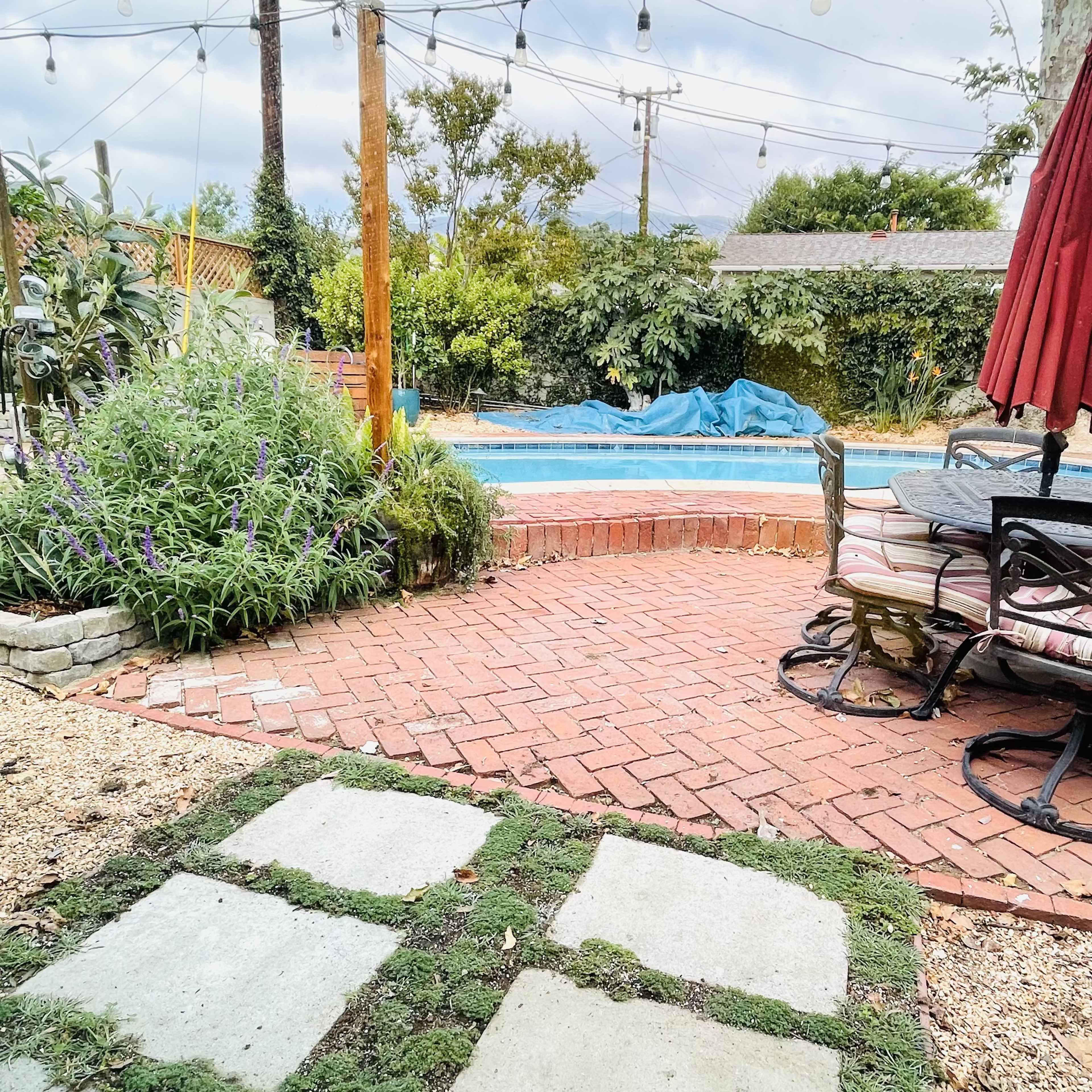 A brick patio with grass insets leads to a swimming pool covered by a blue tarp, surrounded by plants and overhead string lights.