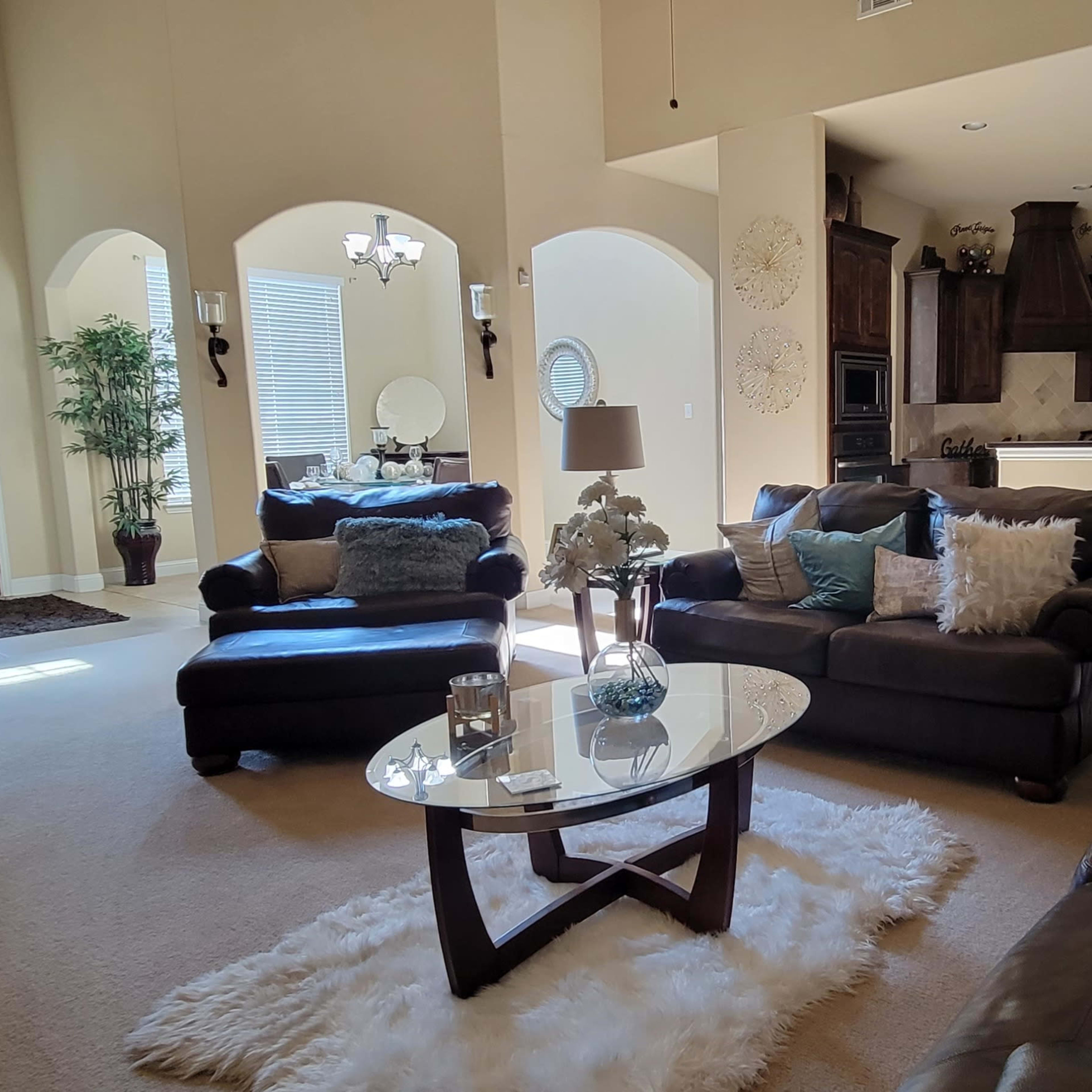 A spacious living room furnished with dark brown sofas, a glass coffee table, and decorative elements, featuring a large window and light-colored walls.