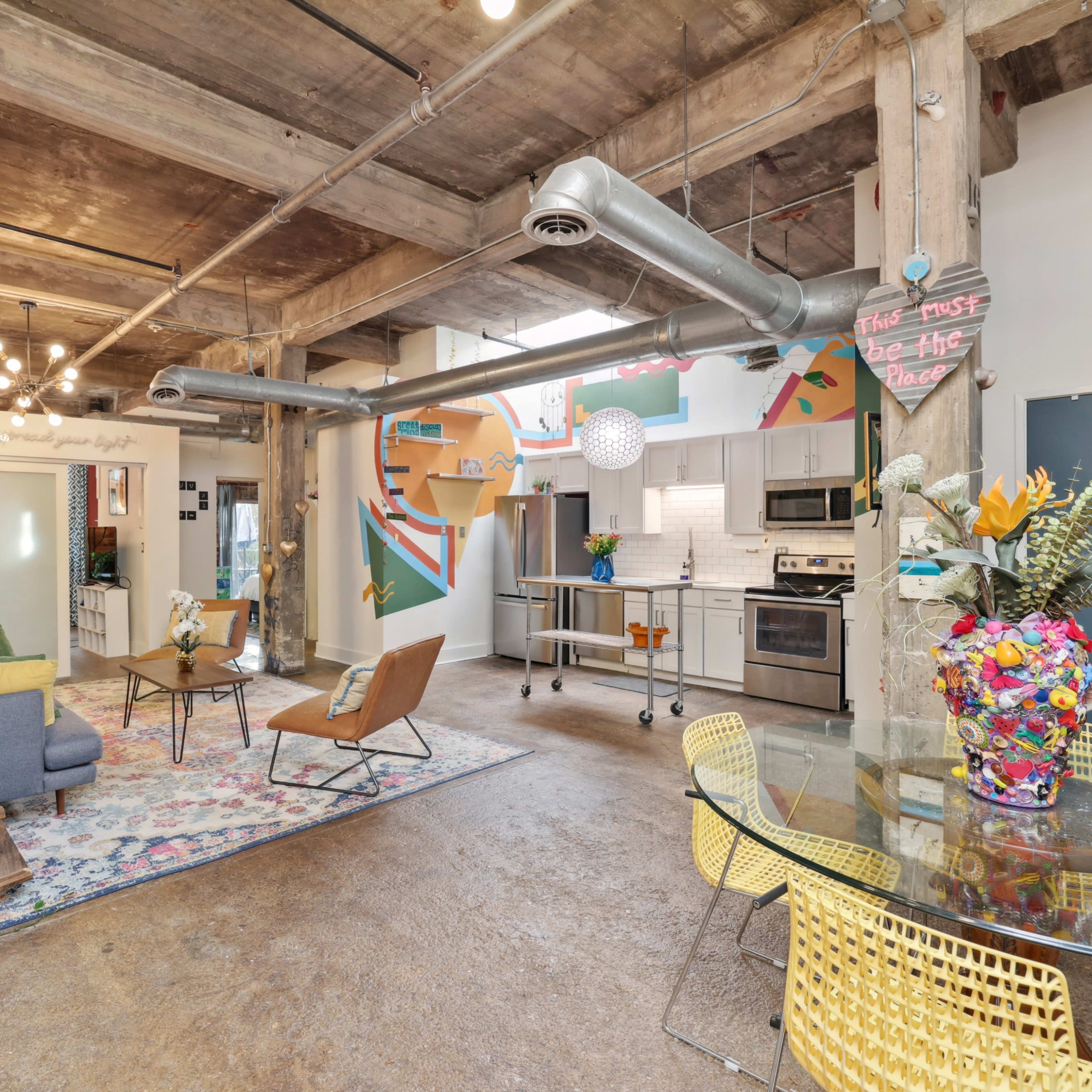 A spacious, urban loft features exposed wooden beams, modern furnishings, and vibrant decorations, including a colorful flower arrangement on a dining table.
