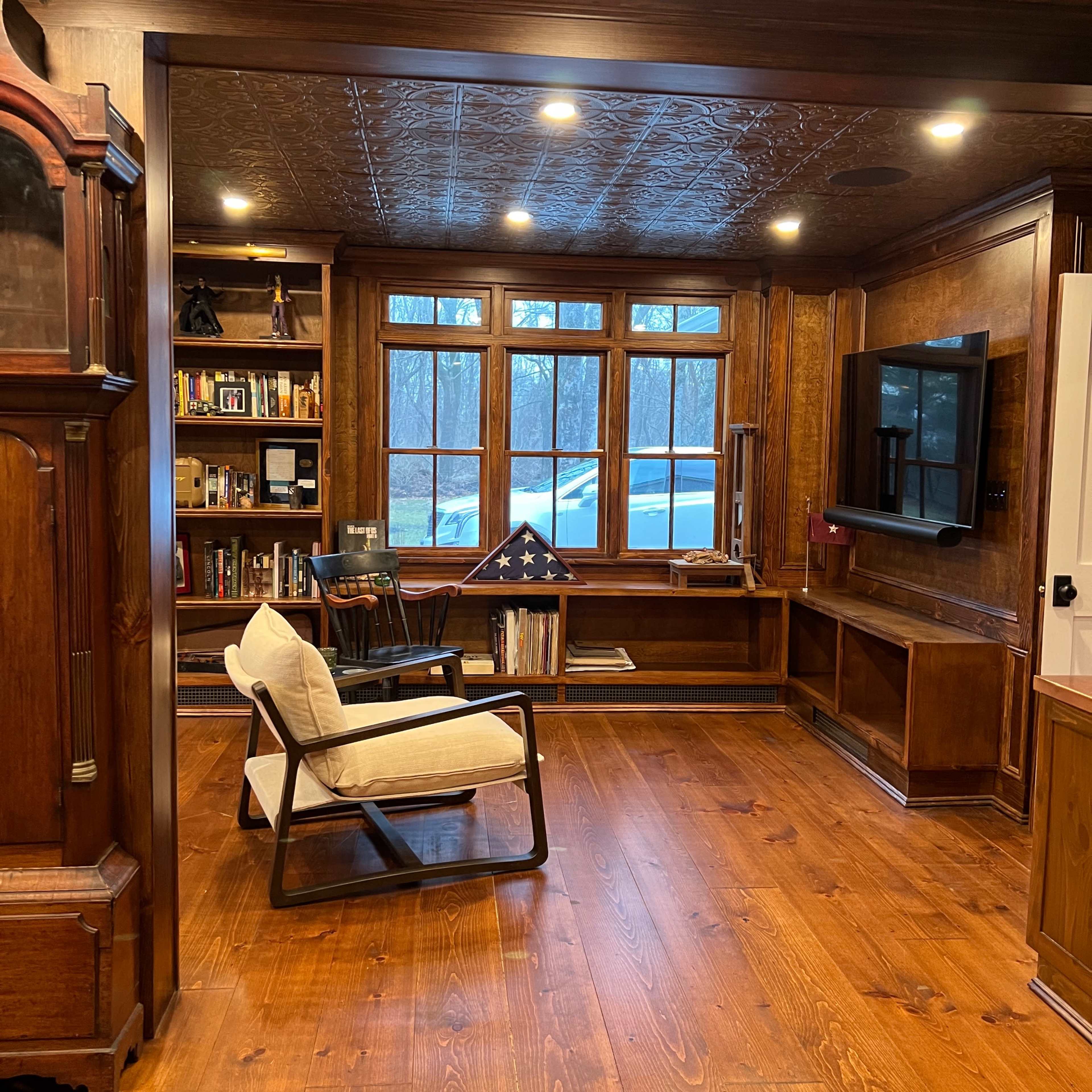 A cozy room with wooden walls, a large window, bookshelves filled with books, a chair, and a television mounted on the wall.