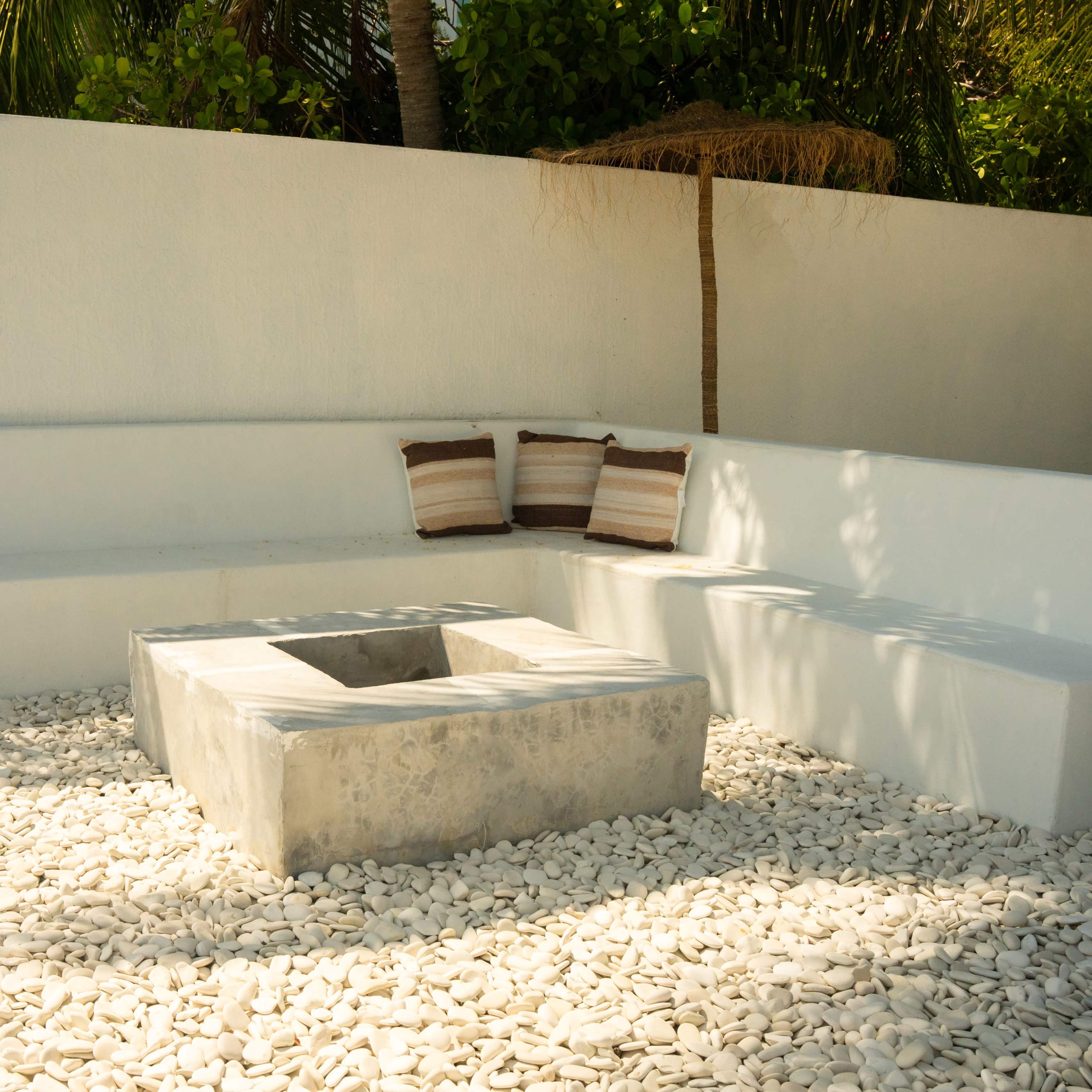 A minimalist outdoor seating area with a concrete fire pit surrounded by white pebbles and low white benches.