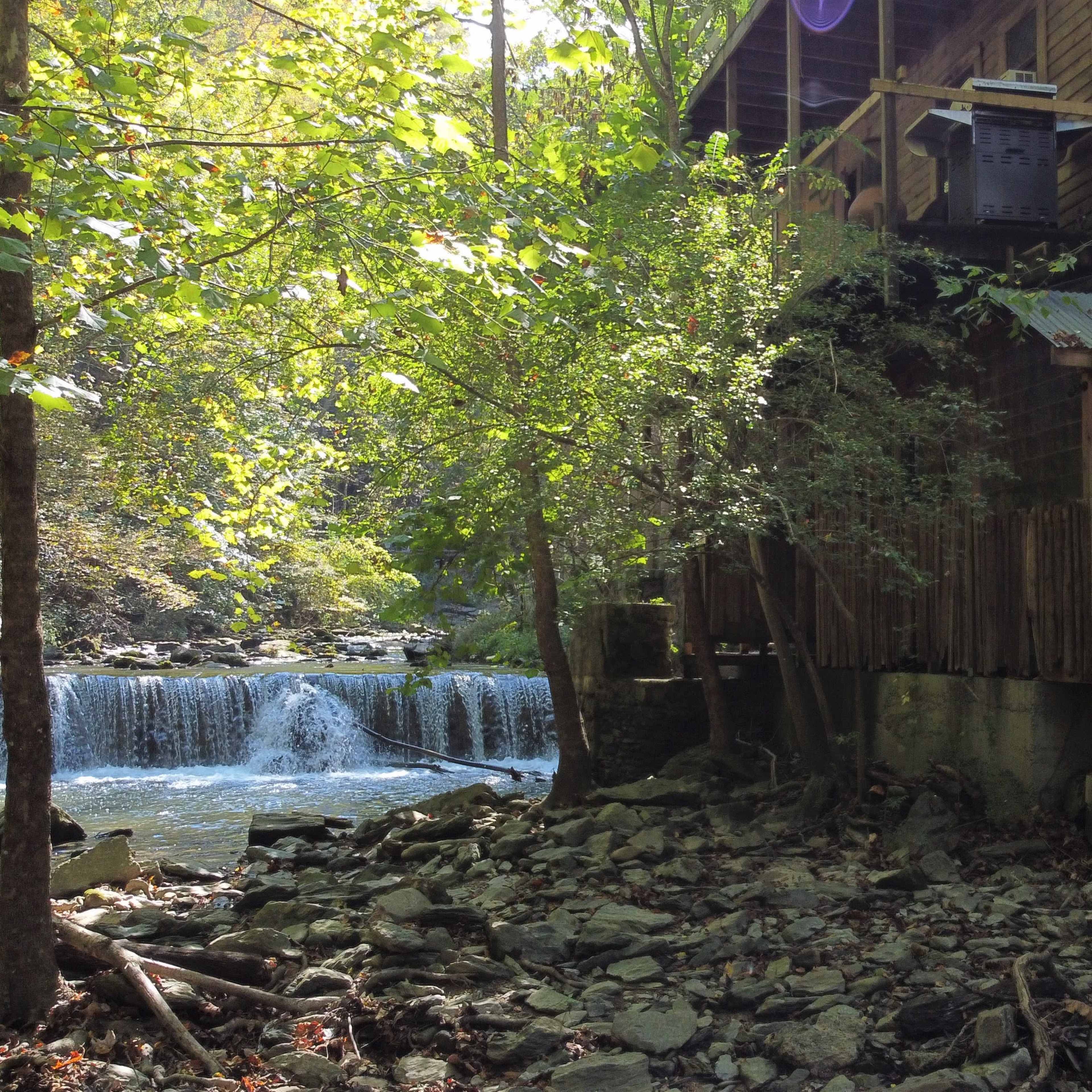 A wooden structure sits alongside a flowing river, with a small waterfall visible in the background amidst lush greenery.