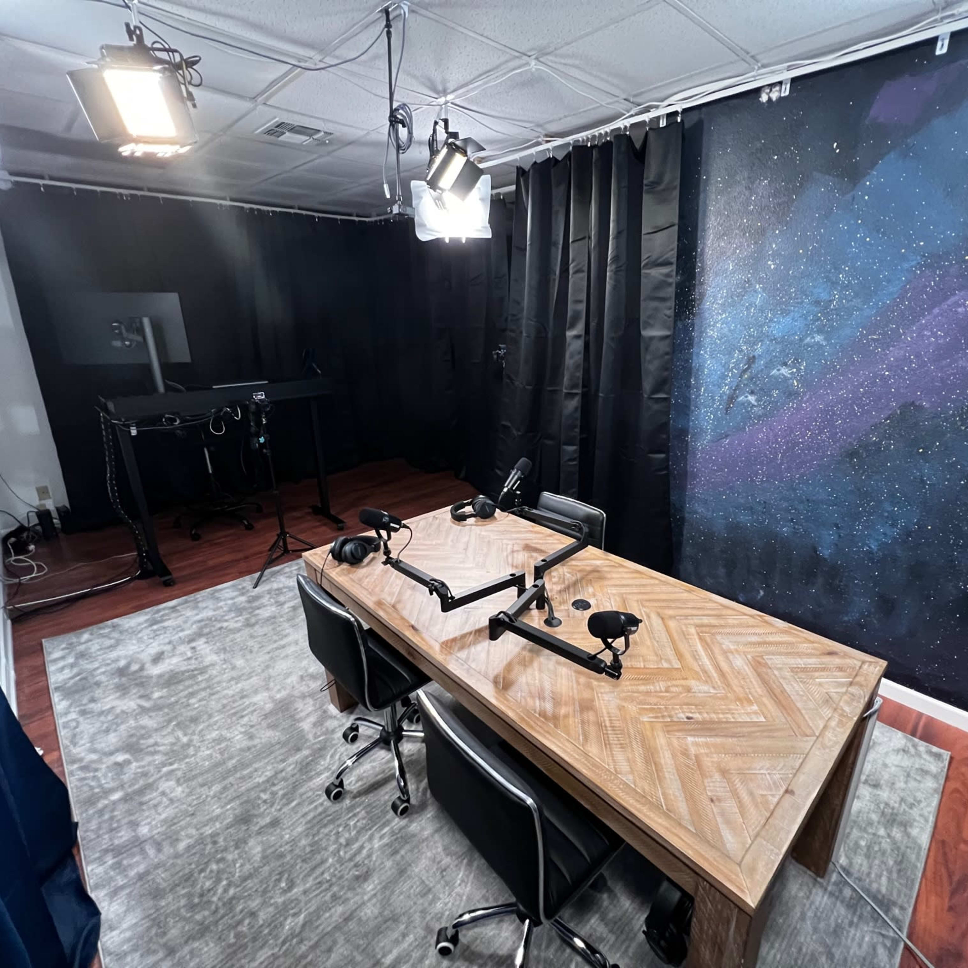 A podcast studio with a wooden table, two microphones on adjustable arms, and a starry backdrop, all under bright studio lights.