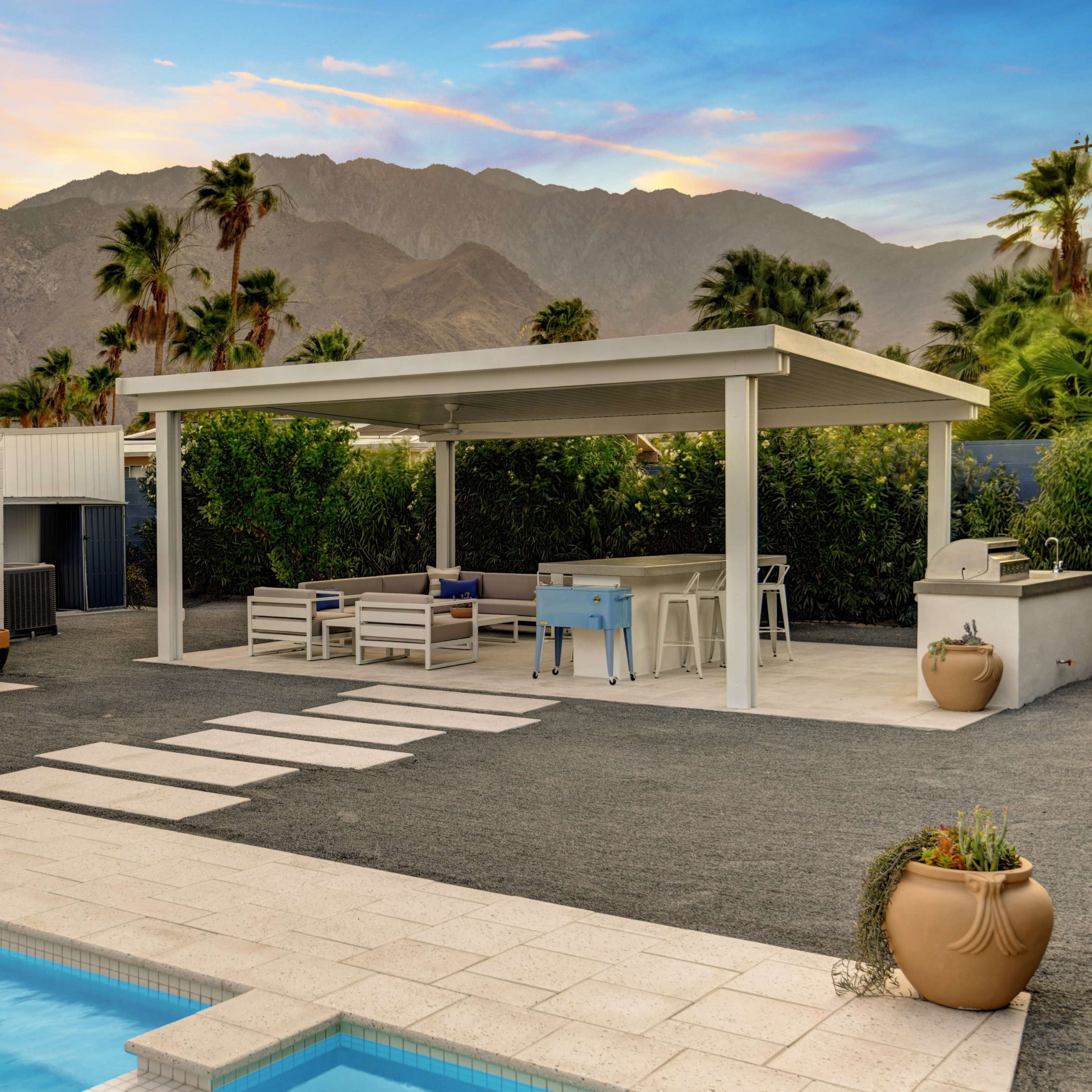 A modern outdoor space featuring a pool, a covered seating area, and a backdrop of mountains under a colorful sky.