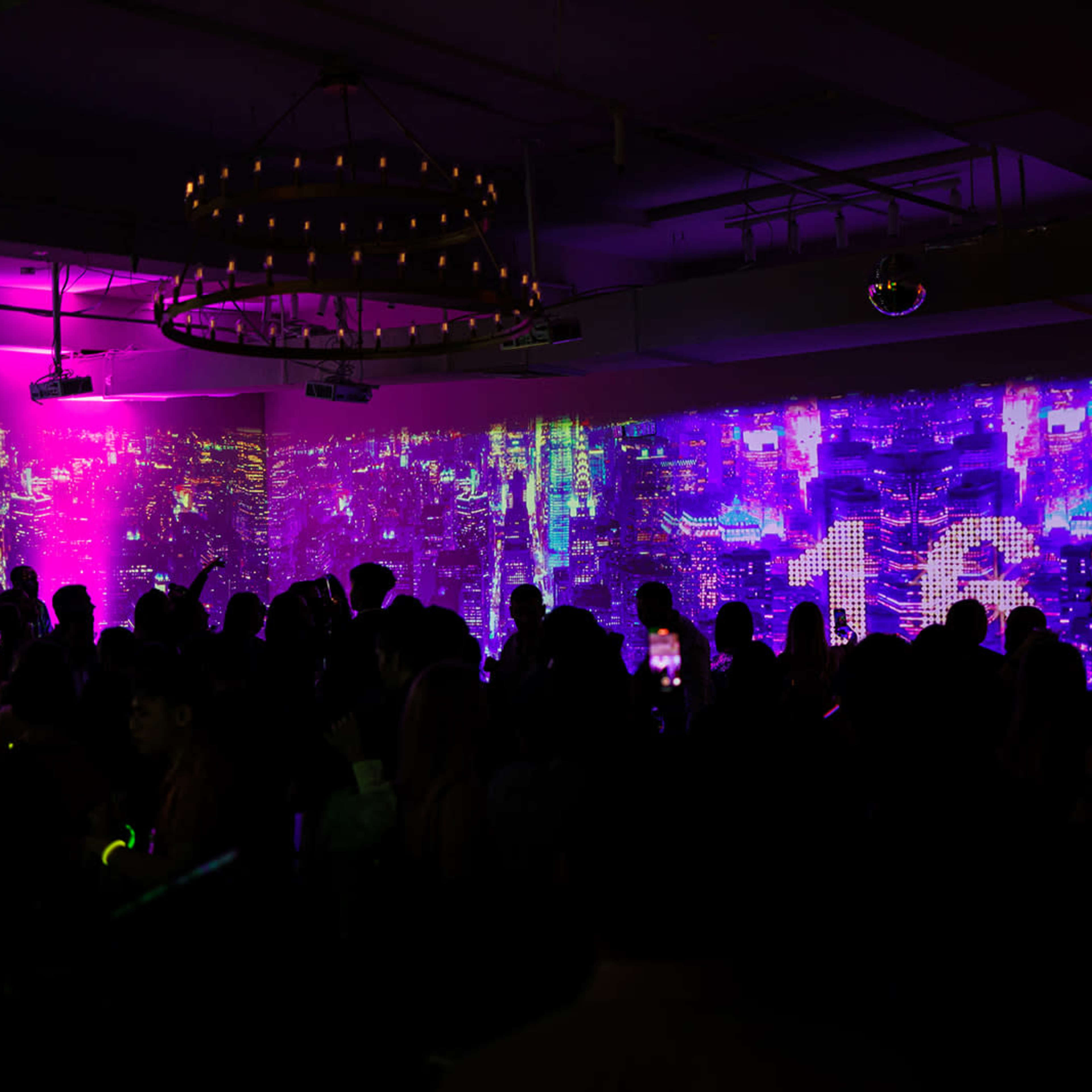 A crowd of people gathers under colorful lights and projected visuals in an indoor venue.