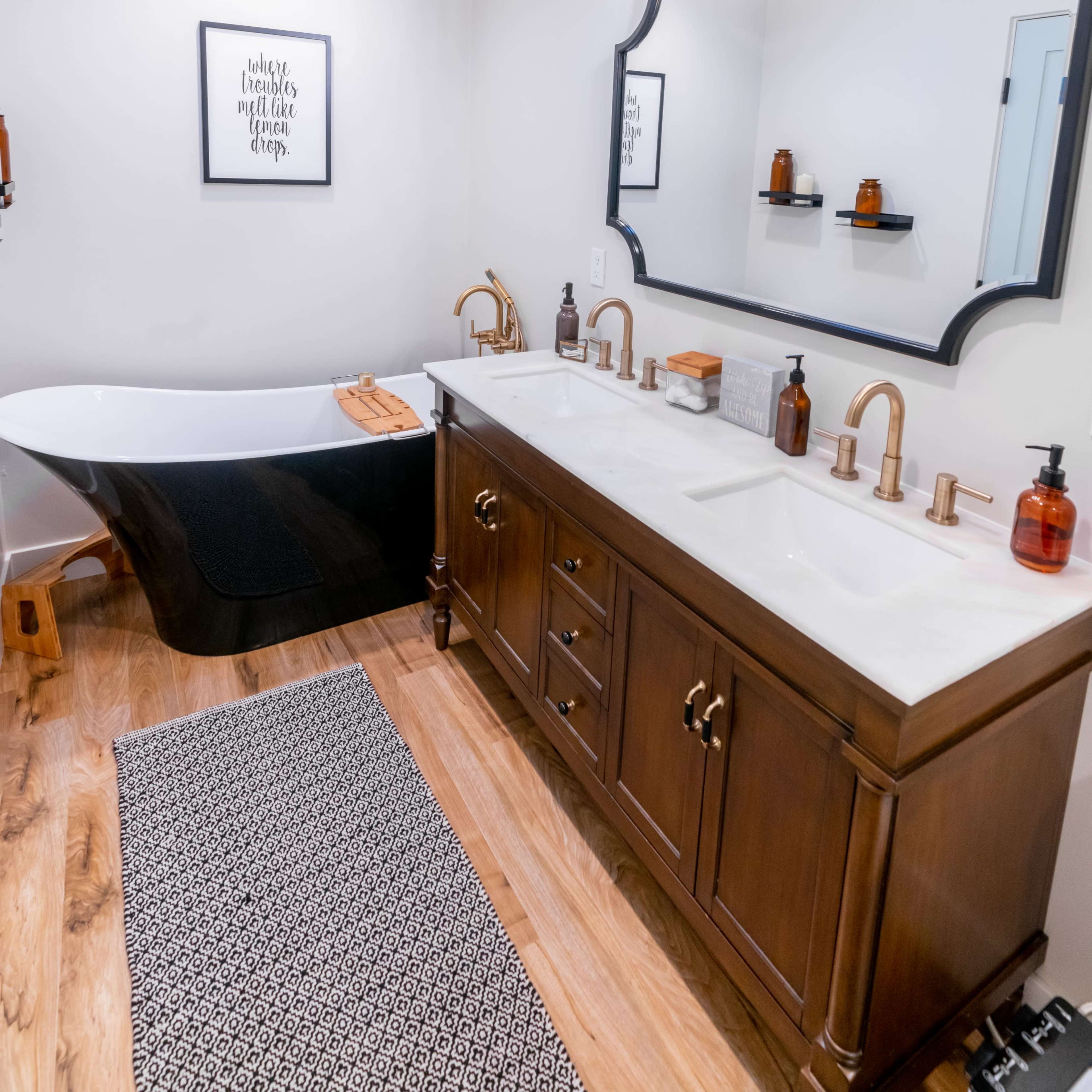 A modern bathroom featuring a freestanding black and white bathtub, a wooden double sink vanity with brass fixtures, and a decorative wall mirror.