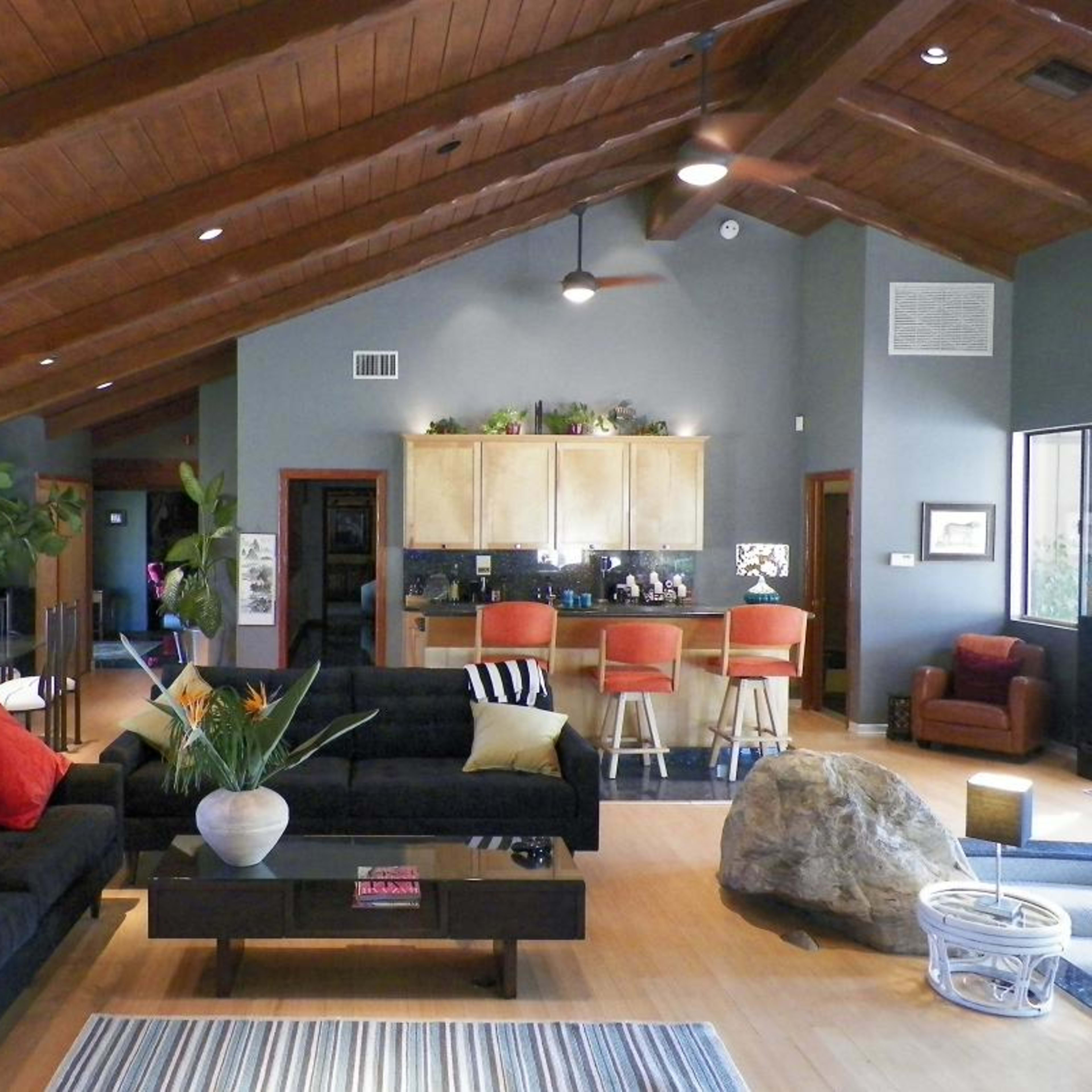 A spacious living area features black sofas with red pillows, a wooden coffee table, and a kitchen bar in the background.