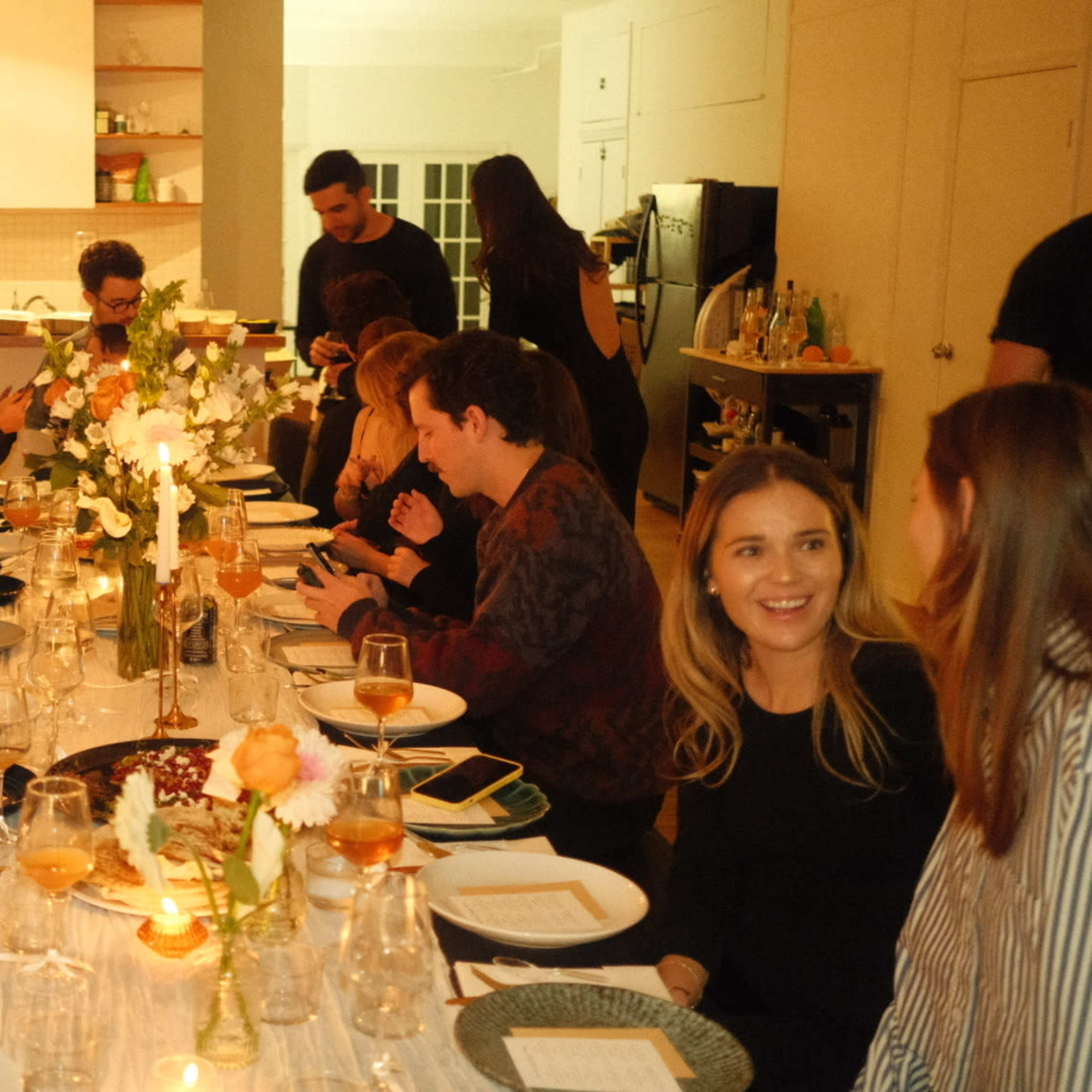 A large group of people sits around a long dining table set with plates and glasses in a warmly lit room.