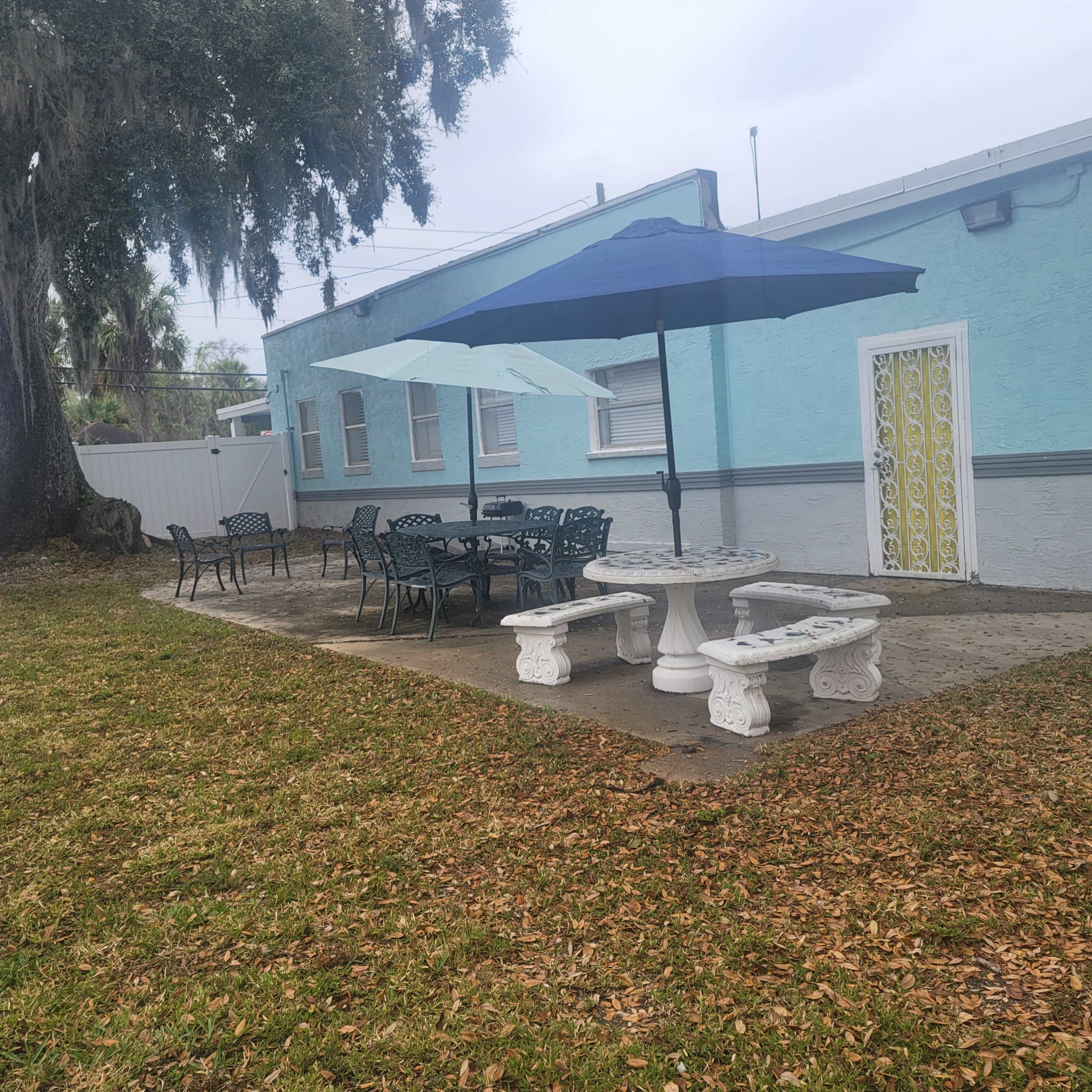 A patio area features two large umbrellas over stone tables and chairs, adjacent to a light blue building.