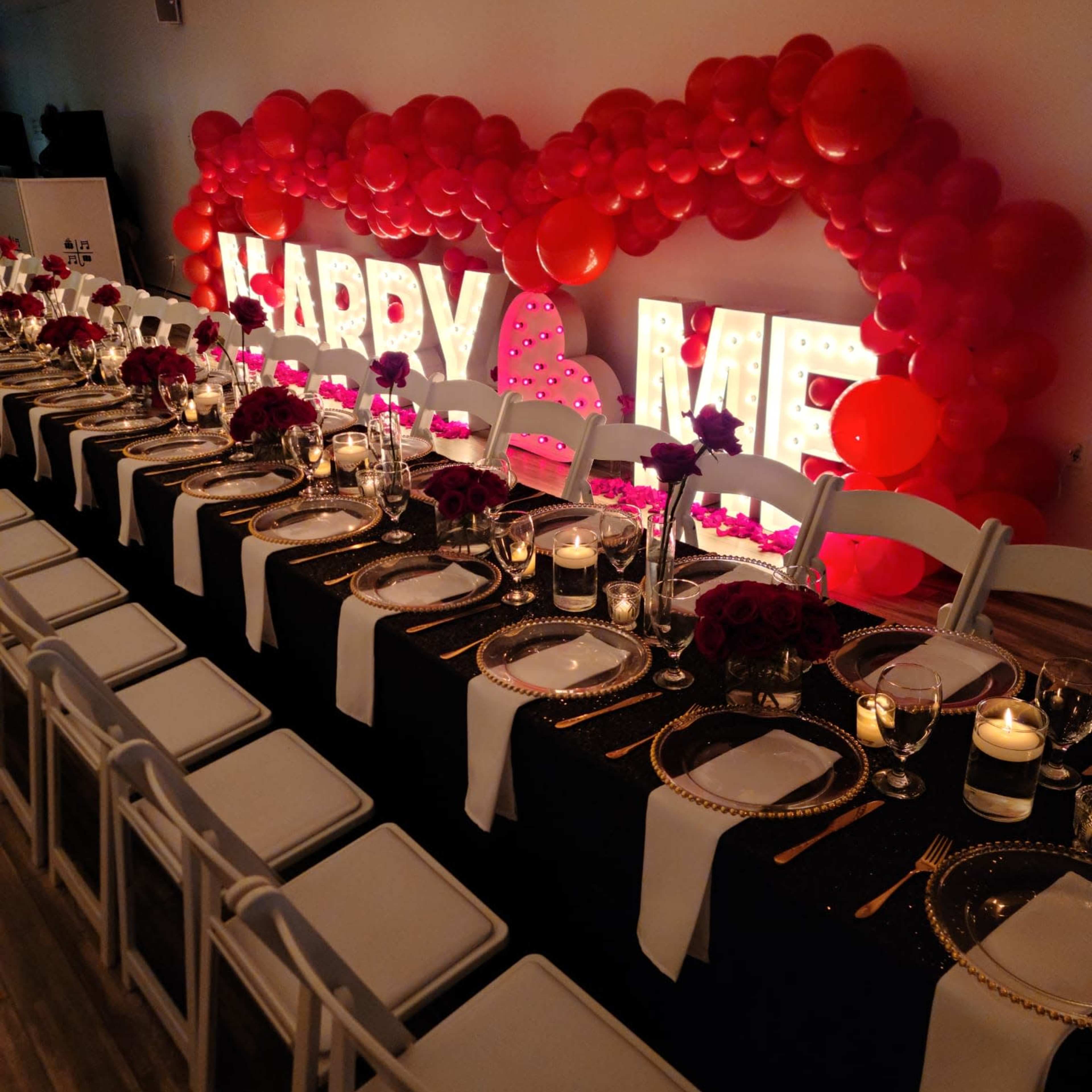 A long dining table is set with elegant tableware, surrounded by white chairs, with a large heart-shaped arrangement of red balloons and illuminated letters spelling "MARRY ME" in the background.