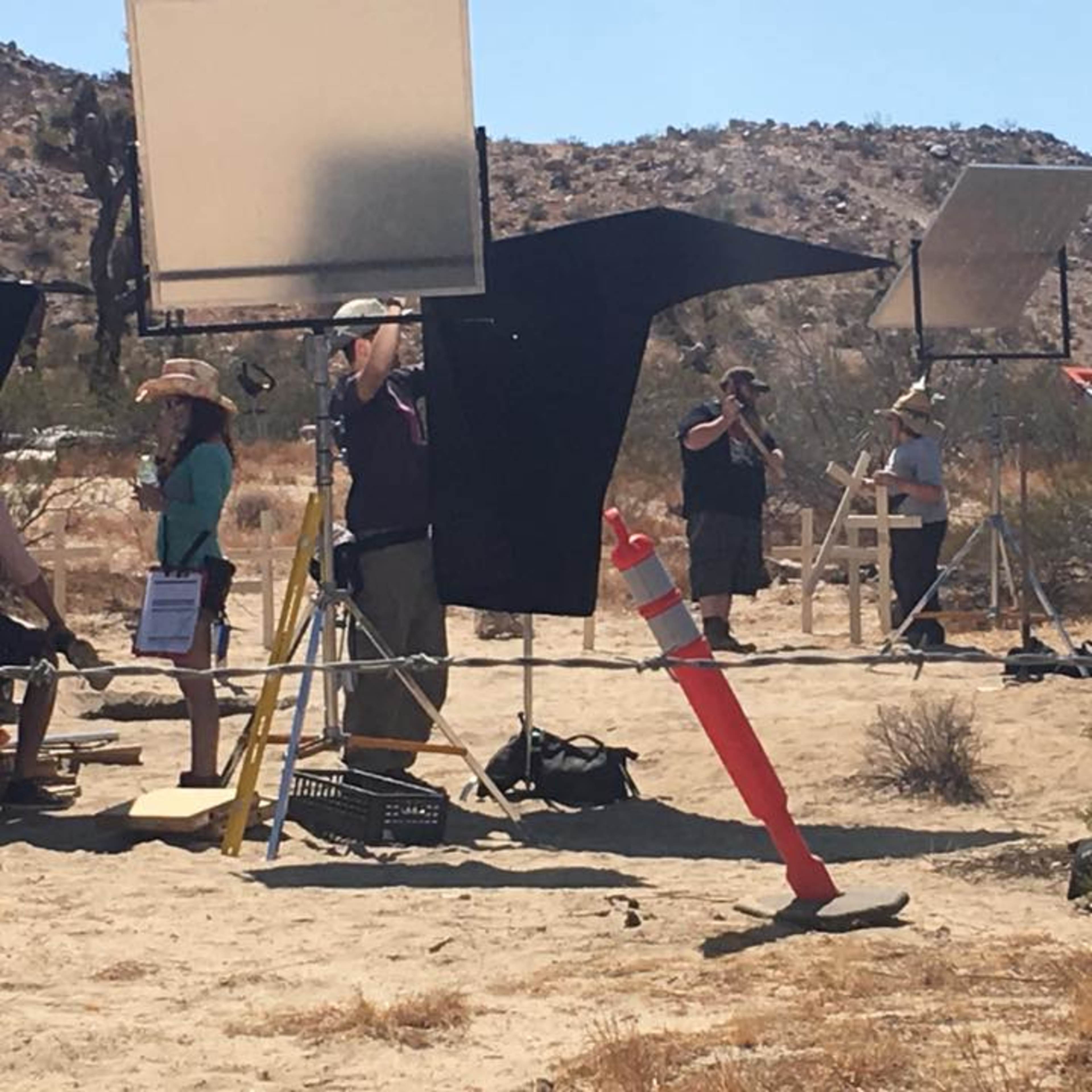A film crew is set up in a desert landscape with various lighting equipment and a wooden structure in the background.