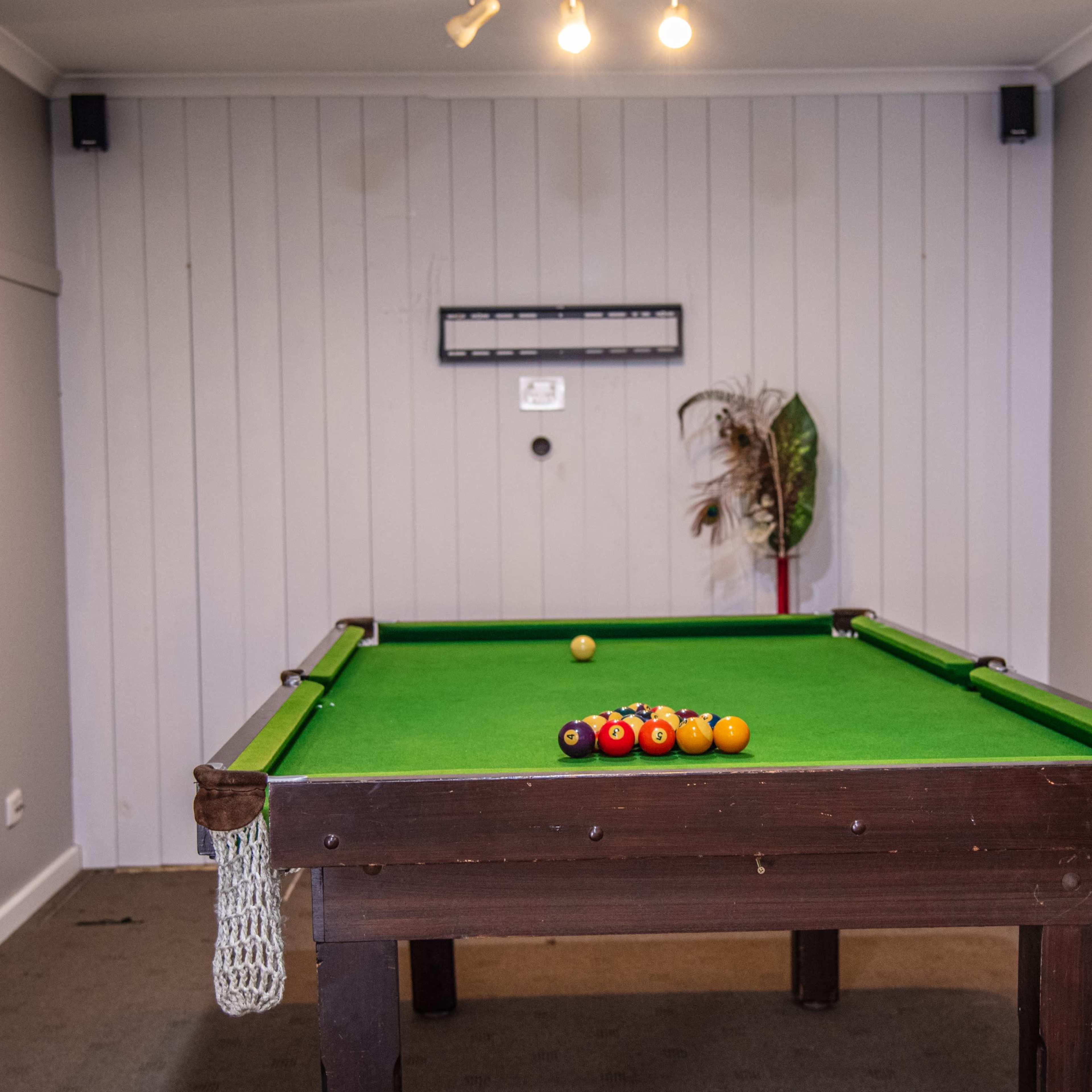 A green billiard table with a set of pool balls arranged on it is positioned in a room featuring pale walls and a small plant in the corner.
