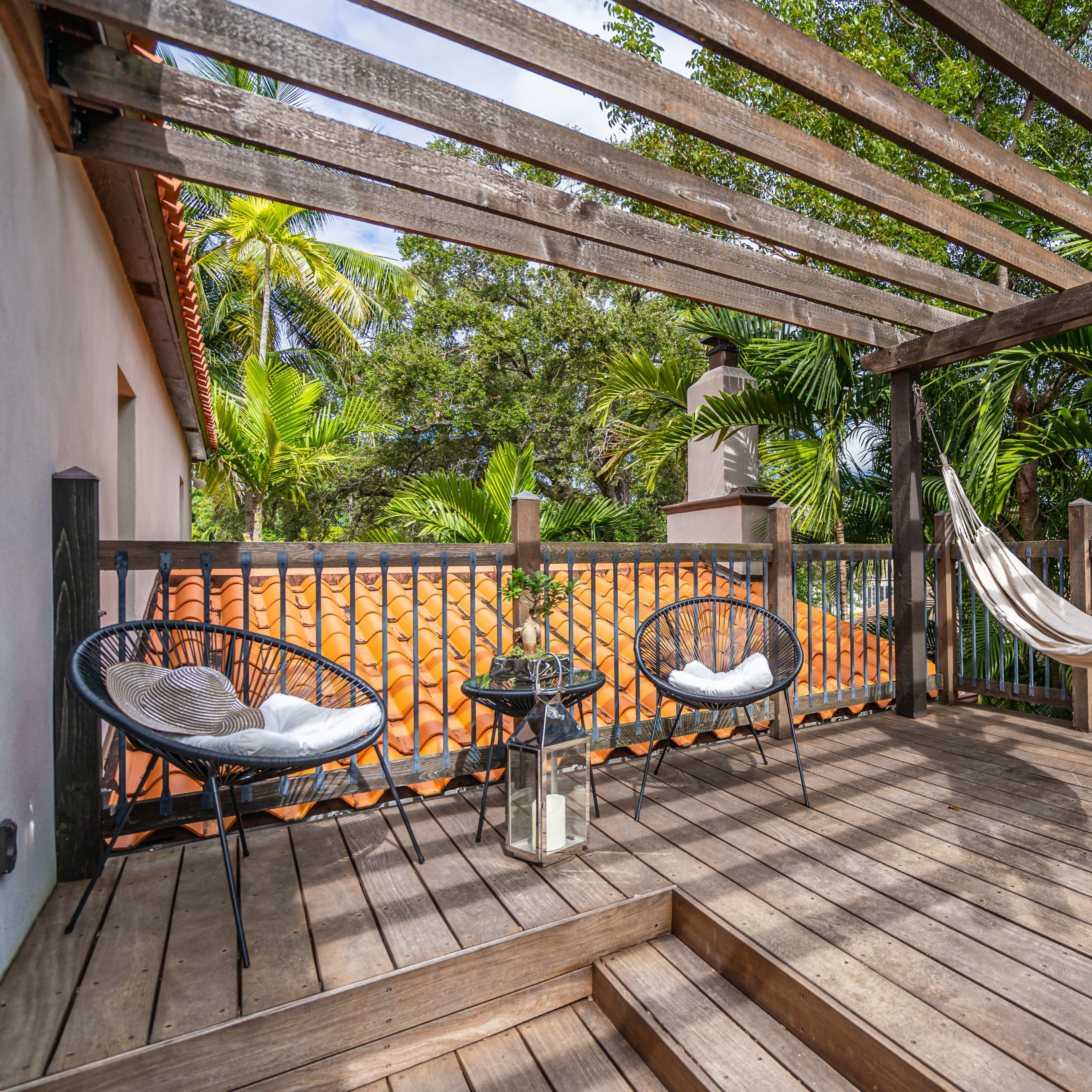 A two-story house with green awnings, surrounded by palm trees, featuring a swimming pool with a brick patio.