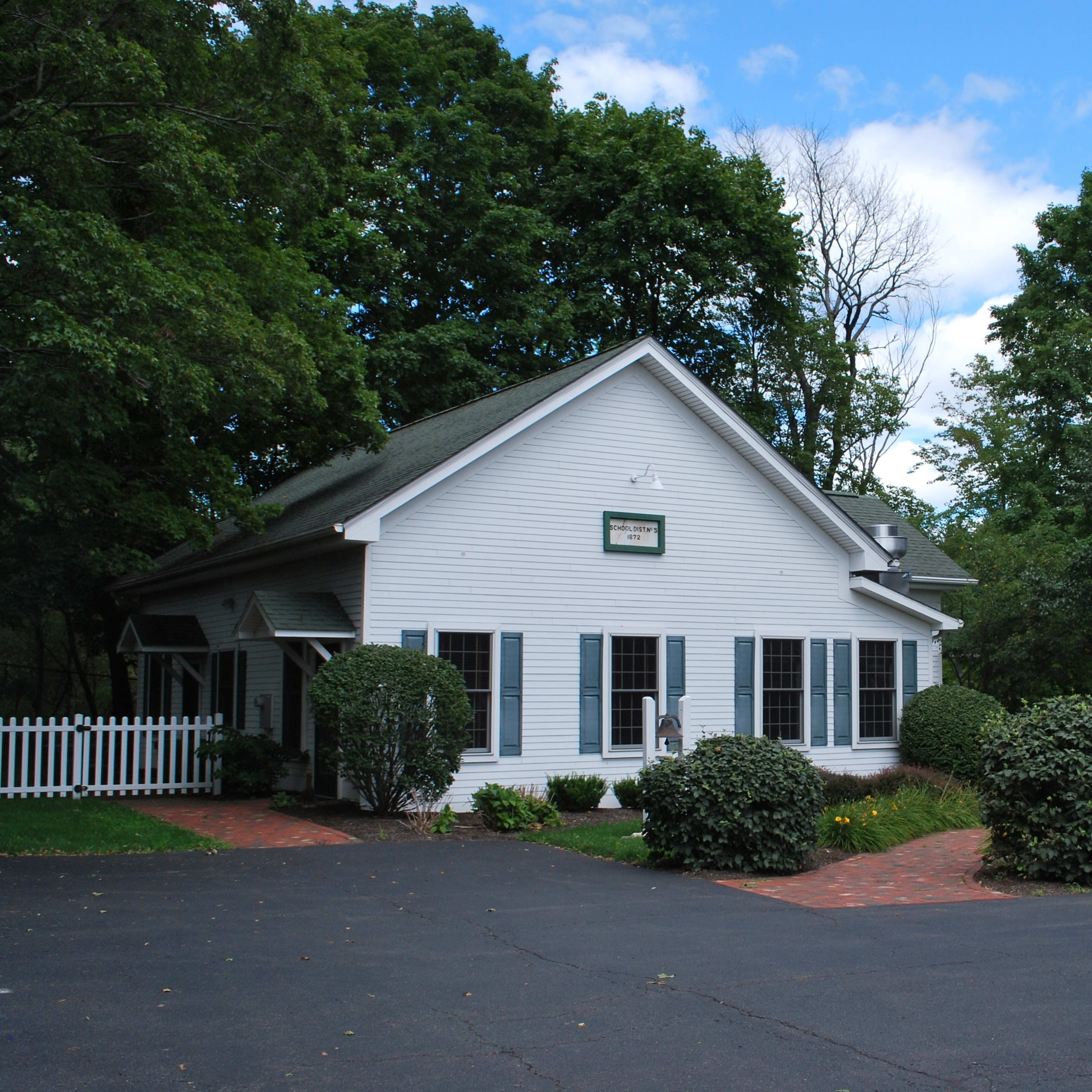 A white house with blue shutters is surrounded by neatly trimmed bushes and trees, with a brick walkway leading to the entrance.