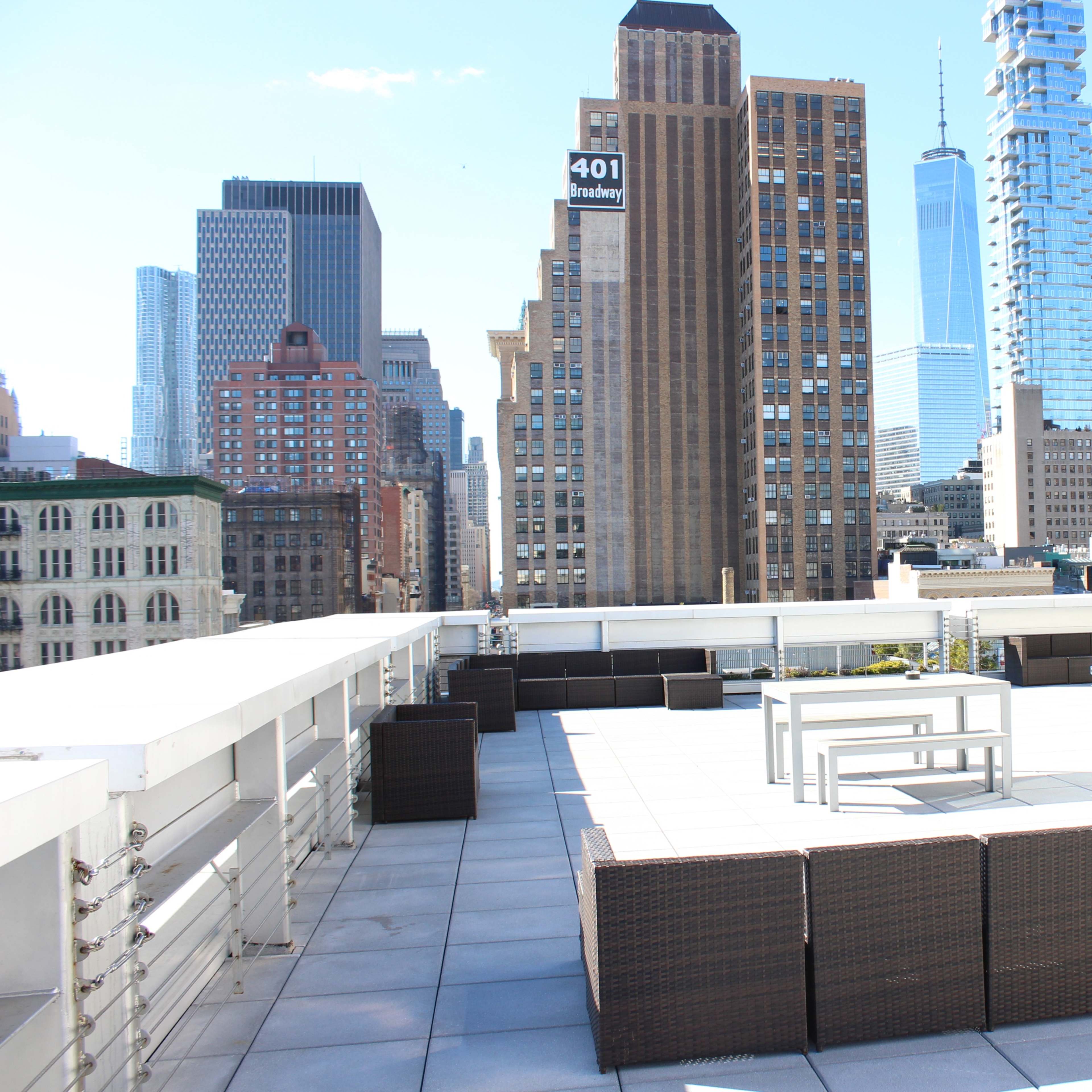 A rooftop terrace features modern furniture with a view of city skyscrapers in a bright, clear sky.