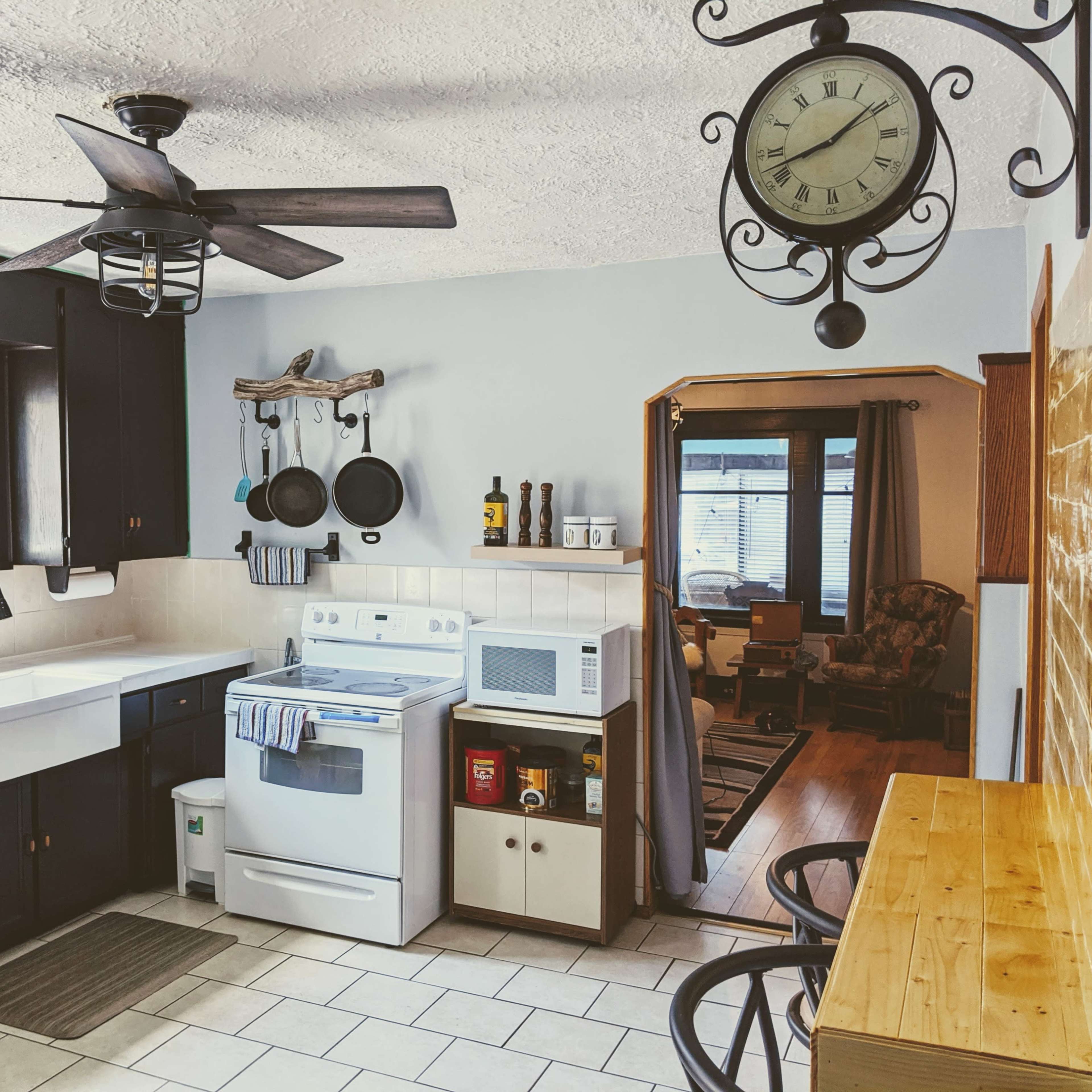 A kitchen with dark cabinets, a white stove, and a countertop equipped with utensils and a microwave, while a clock hangs above and a doorway leads to another room.