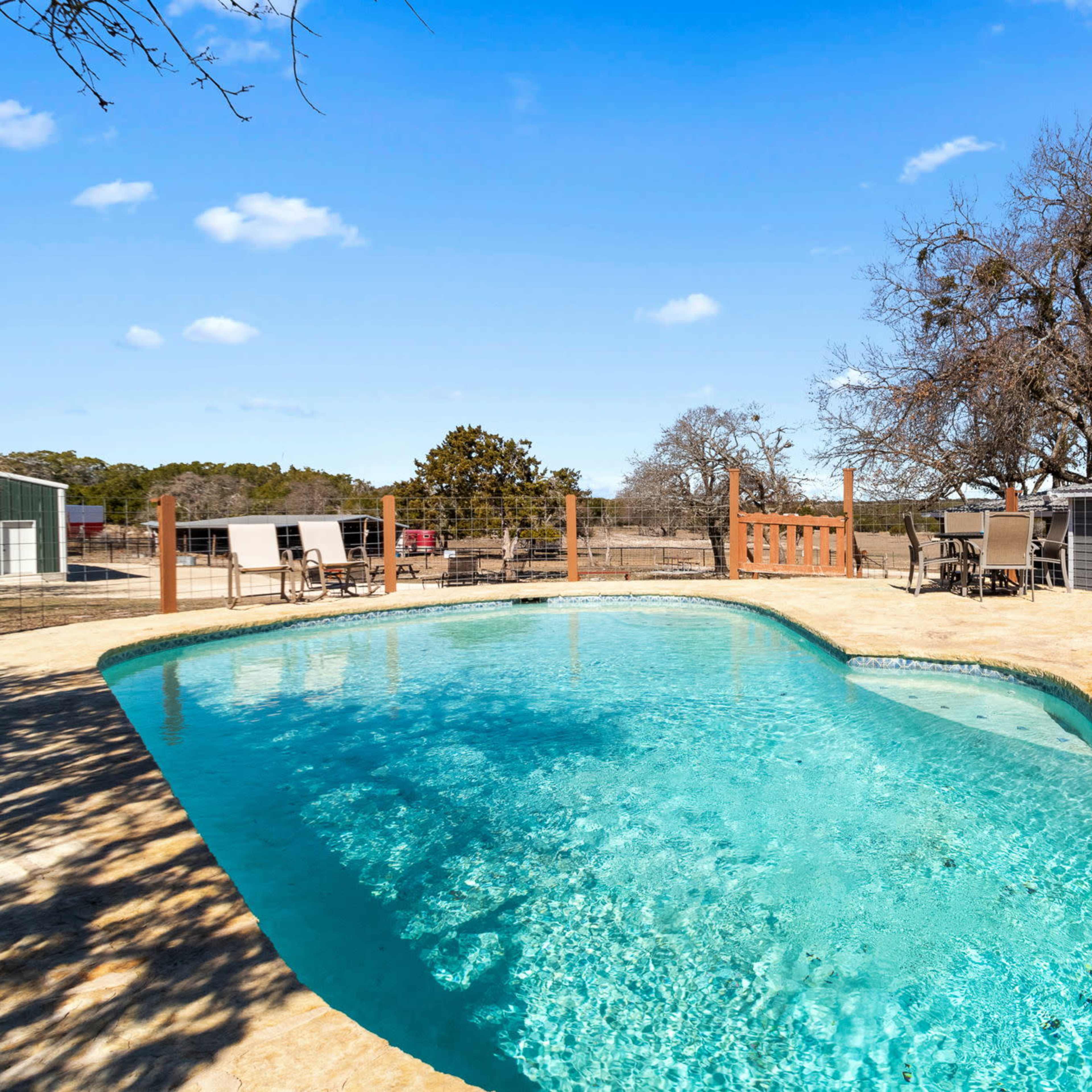 A swimming pool surrounded by outdoor seating and a wooden fence, with trees and a grey building in the background.