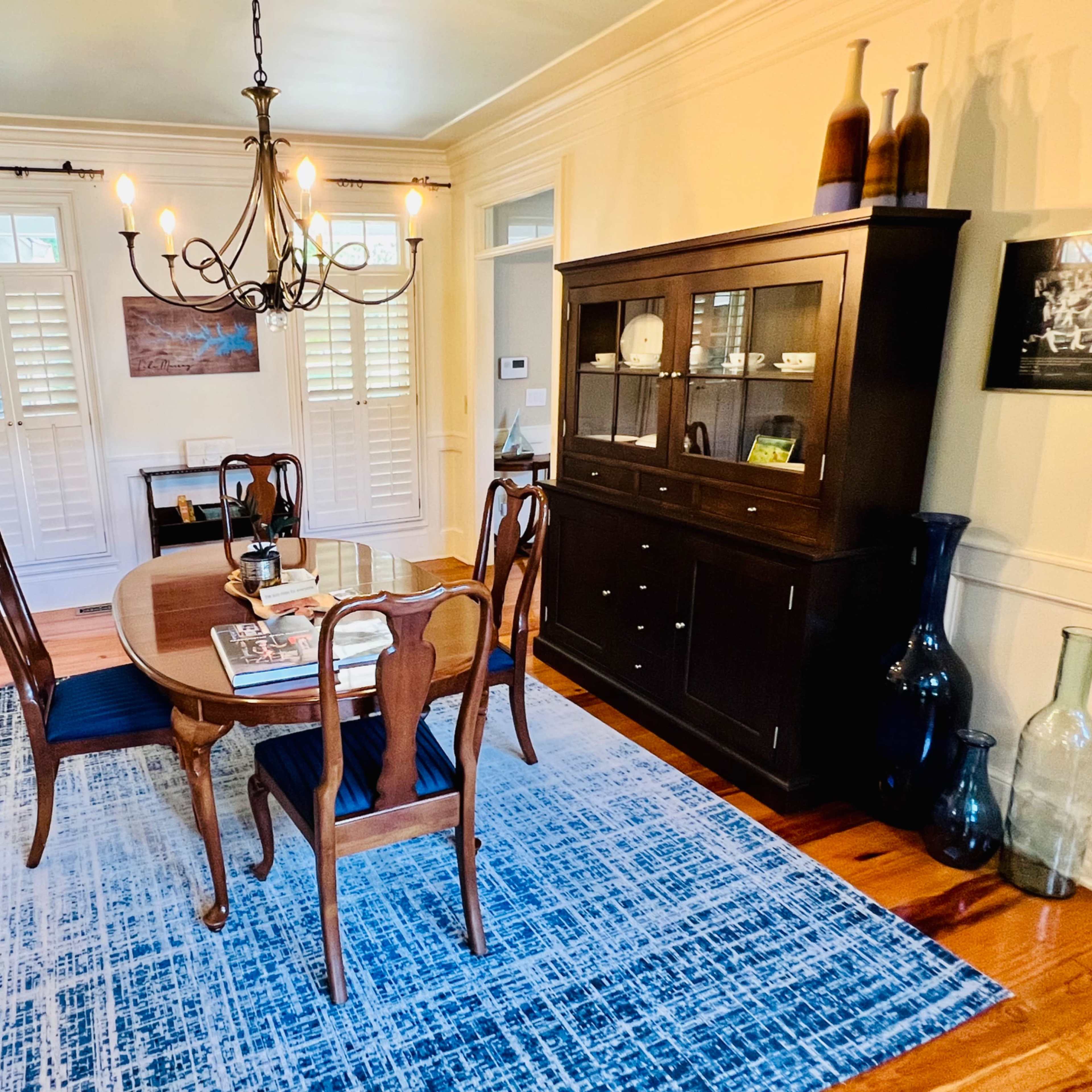 A dining room features a wooden table surrounded by four chairs, a chandelier overhead, and a cabinet displaying various items, with blue and green vases on the floor.