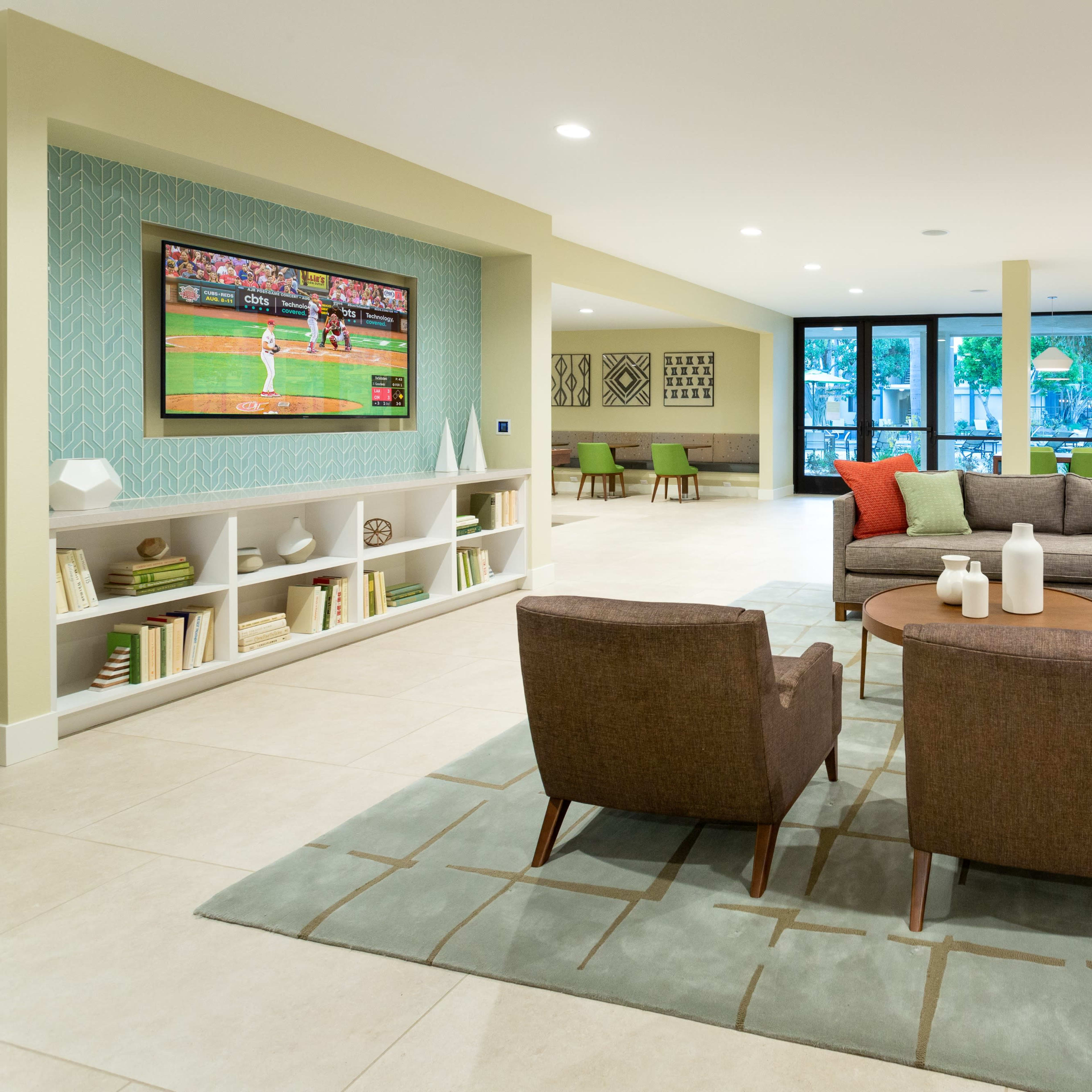 A modern living room features a sectional sofa with colorful cushions, two armchairs, a coffee table, and a TV displaying a baseball game, all set on a tiled floor.