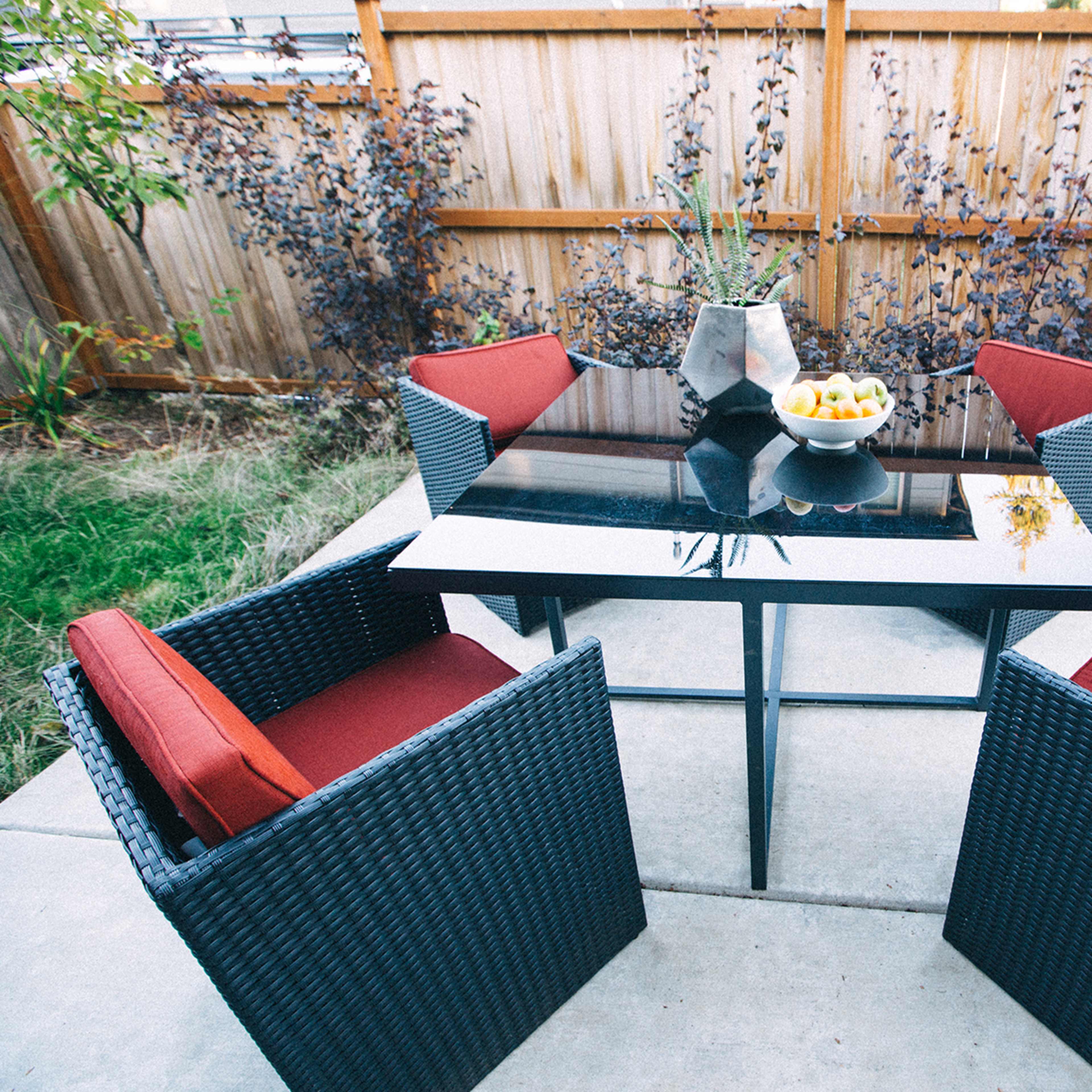 A square glass table surrounded by four wicker chairs with red cushions is set on a concrete patio, with a bowl of fruit on the table and a wooden fence in the background.