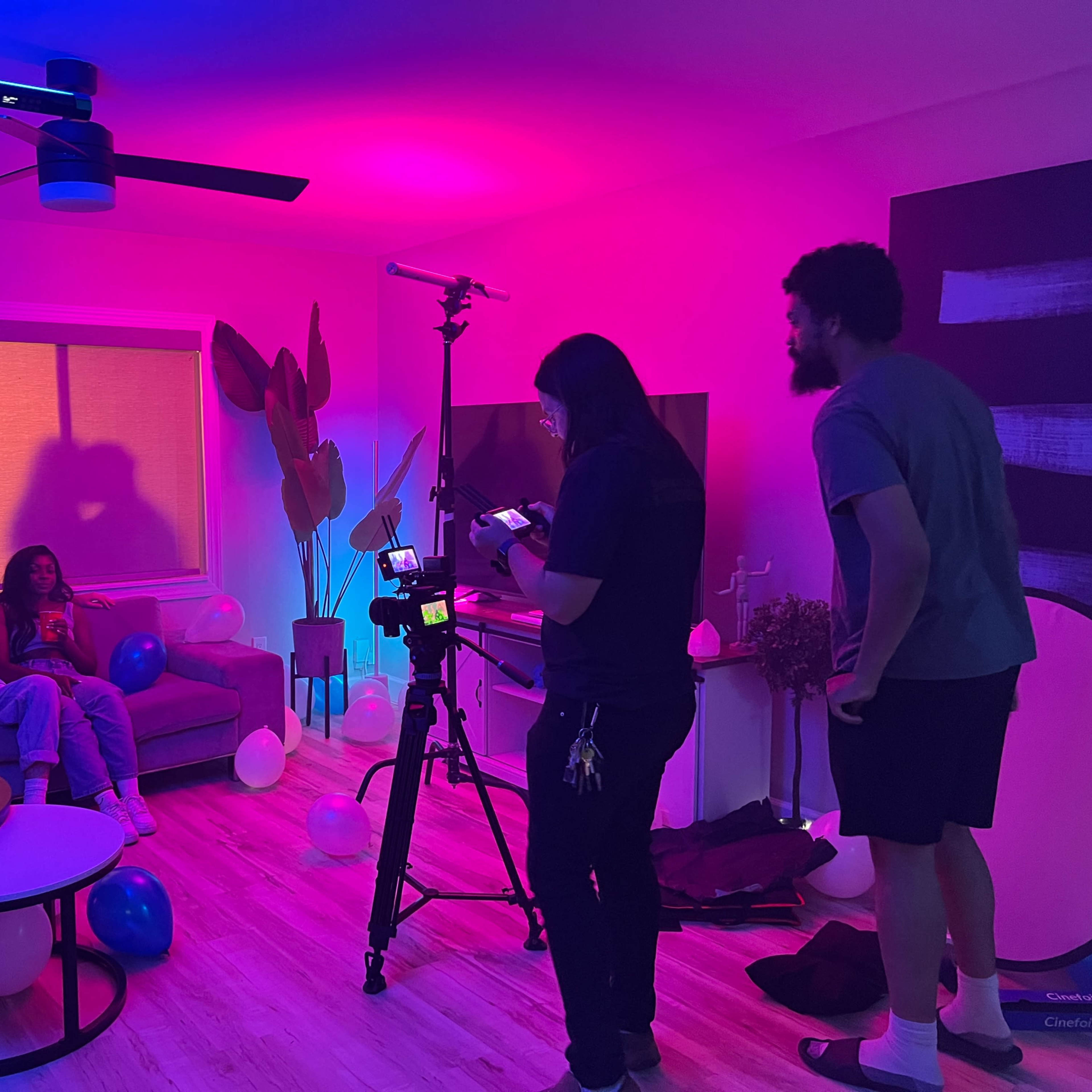A film crew sets up a scene with colorful lighting while two people pose on a couch surrounded by balloons.