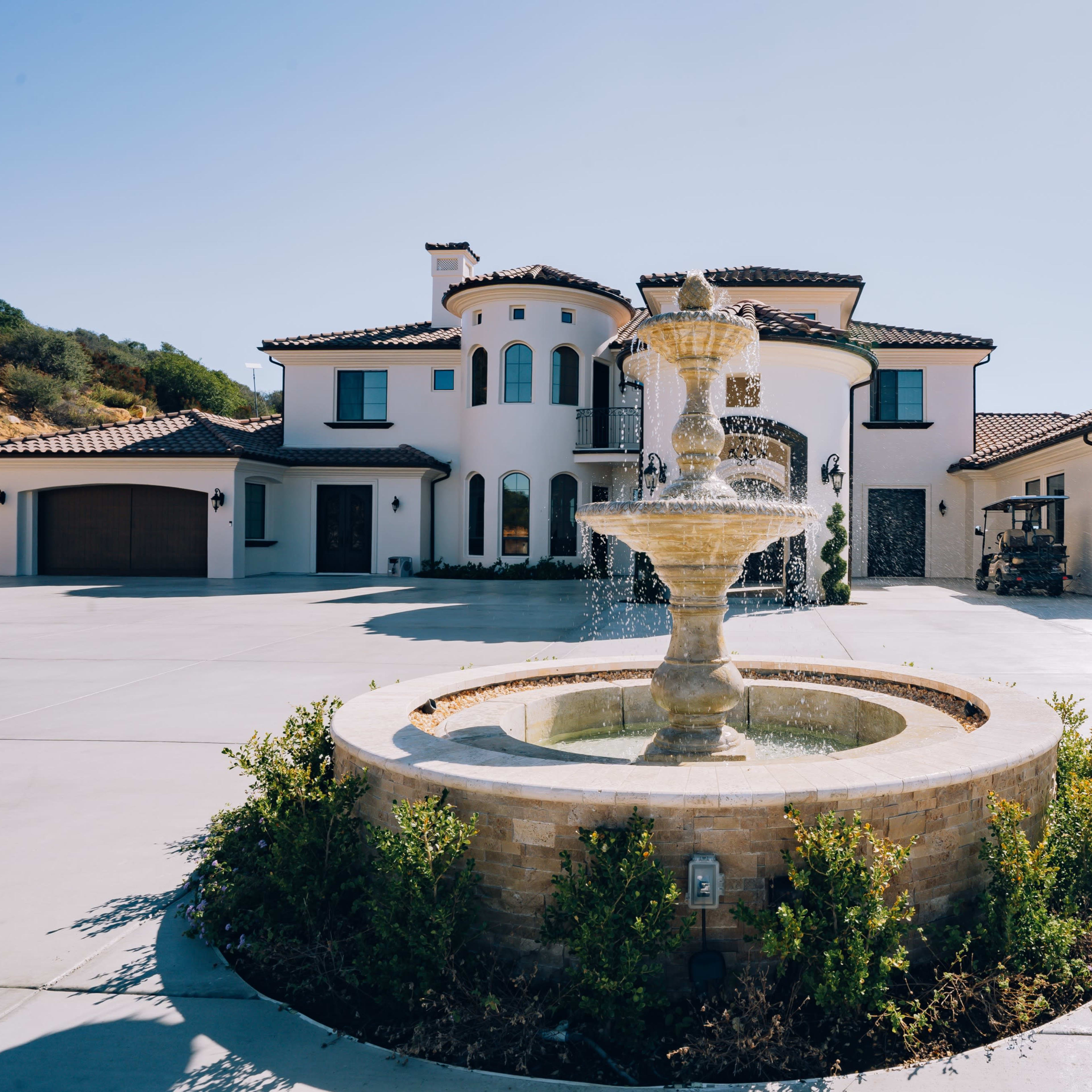 A large fountain is situated in the center of a spacious driveway in front of a modern two-story house with a Mediterranean architectural style.