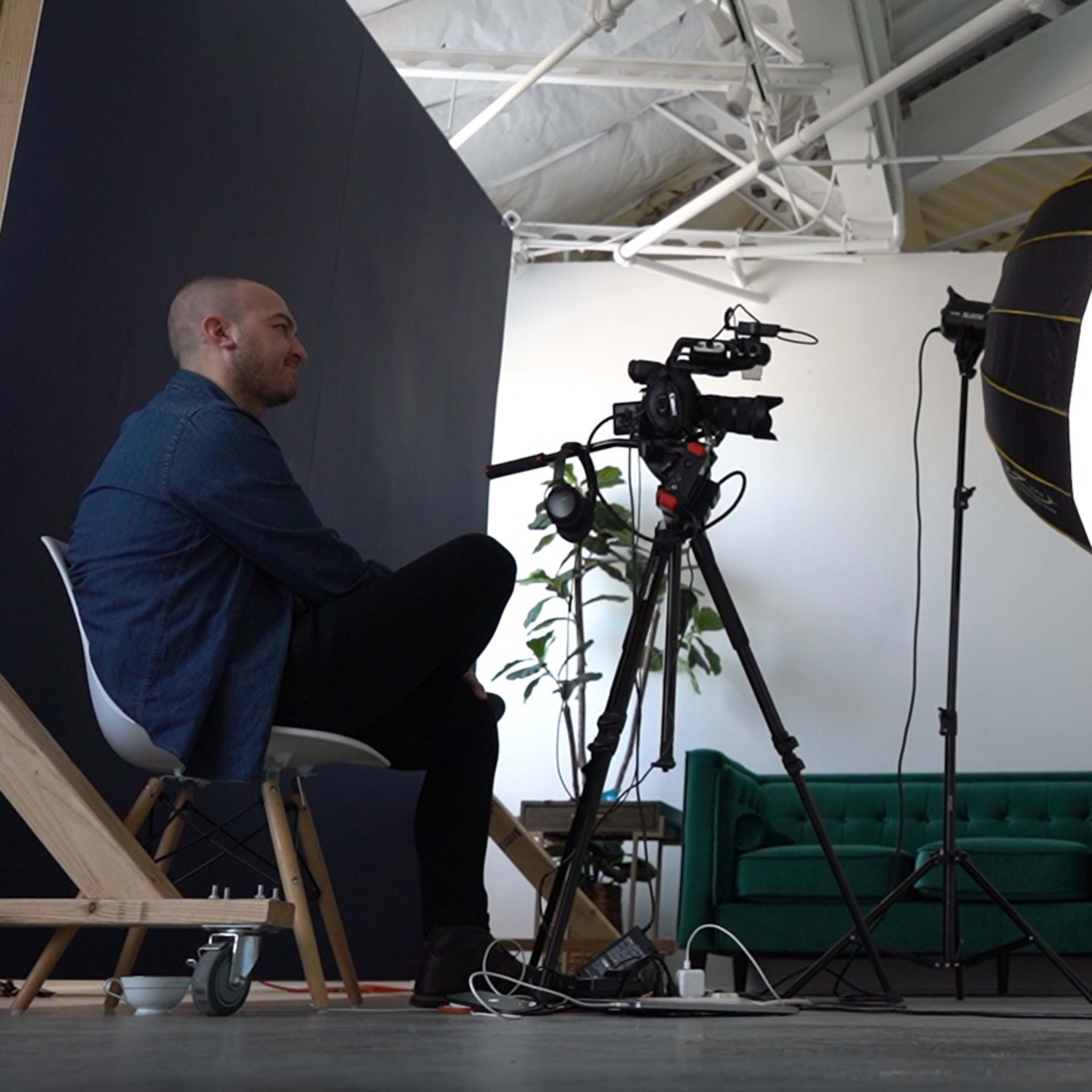 A man and a woman are seated in a well-lit studio, with a camera on a tripod focused on the woman, who is speaking while the man listens.