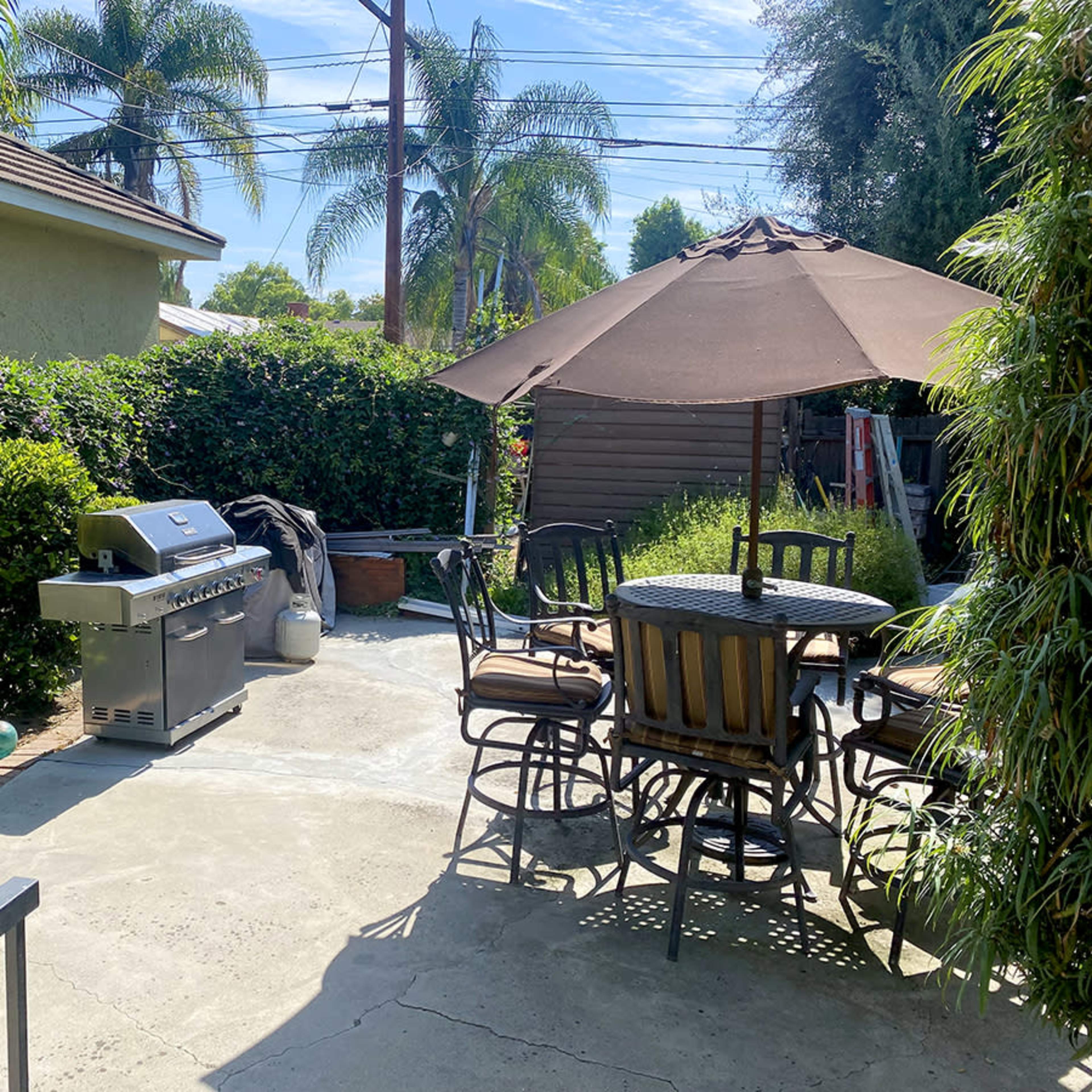 A backyard patio features a grill, a dining set with an umbrella, and surrounding greenery.
