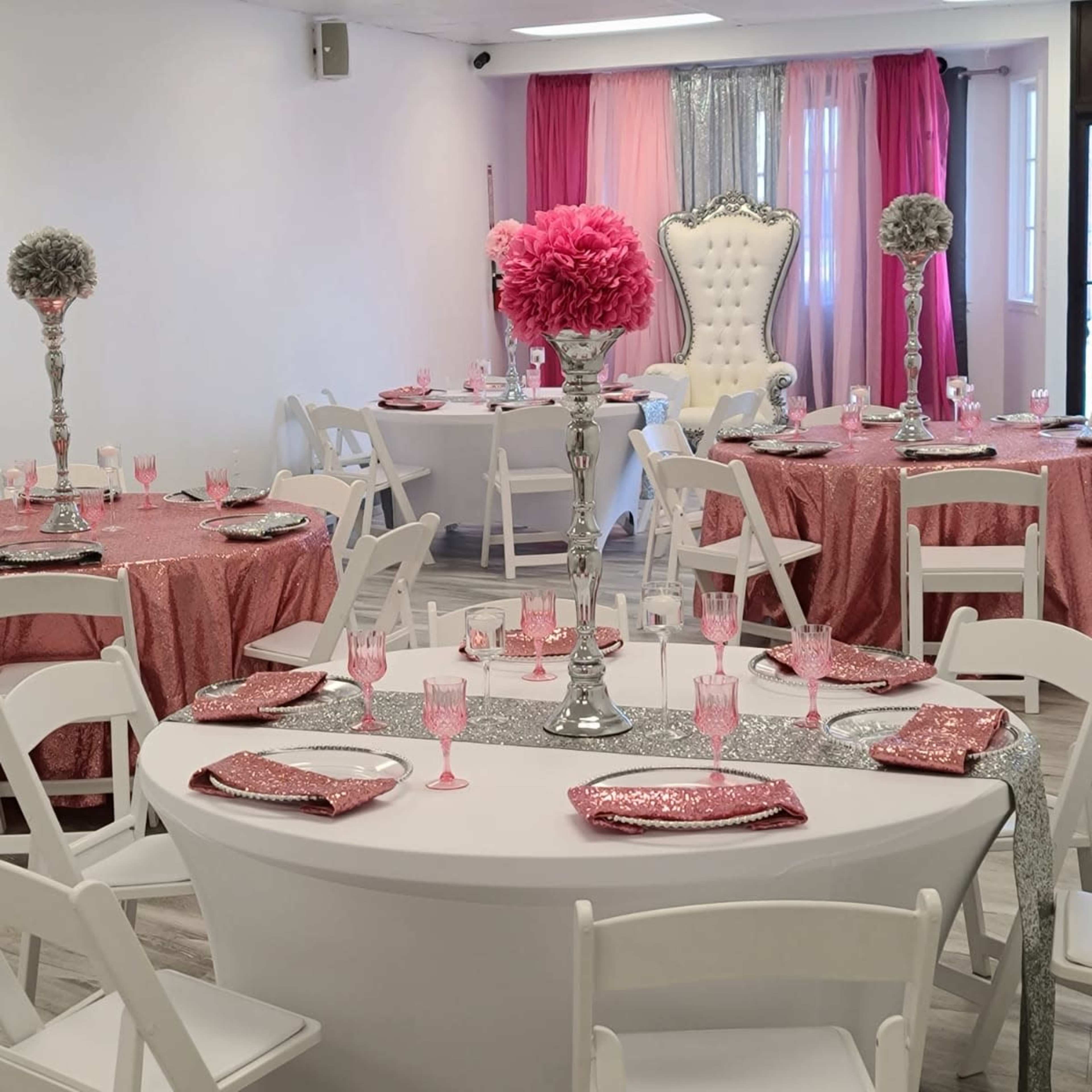 A banquet hall setup with round tables dressed in pink and silver tablecloths, each featuring tall floral centerpieces and elegant tableware.