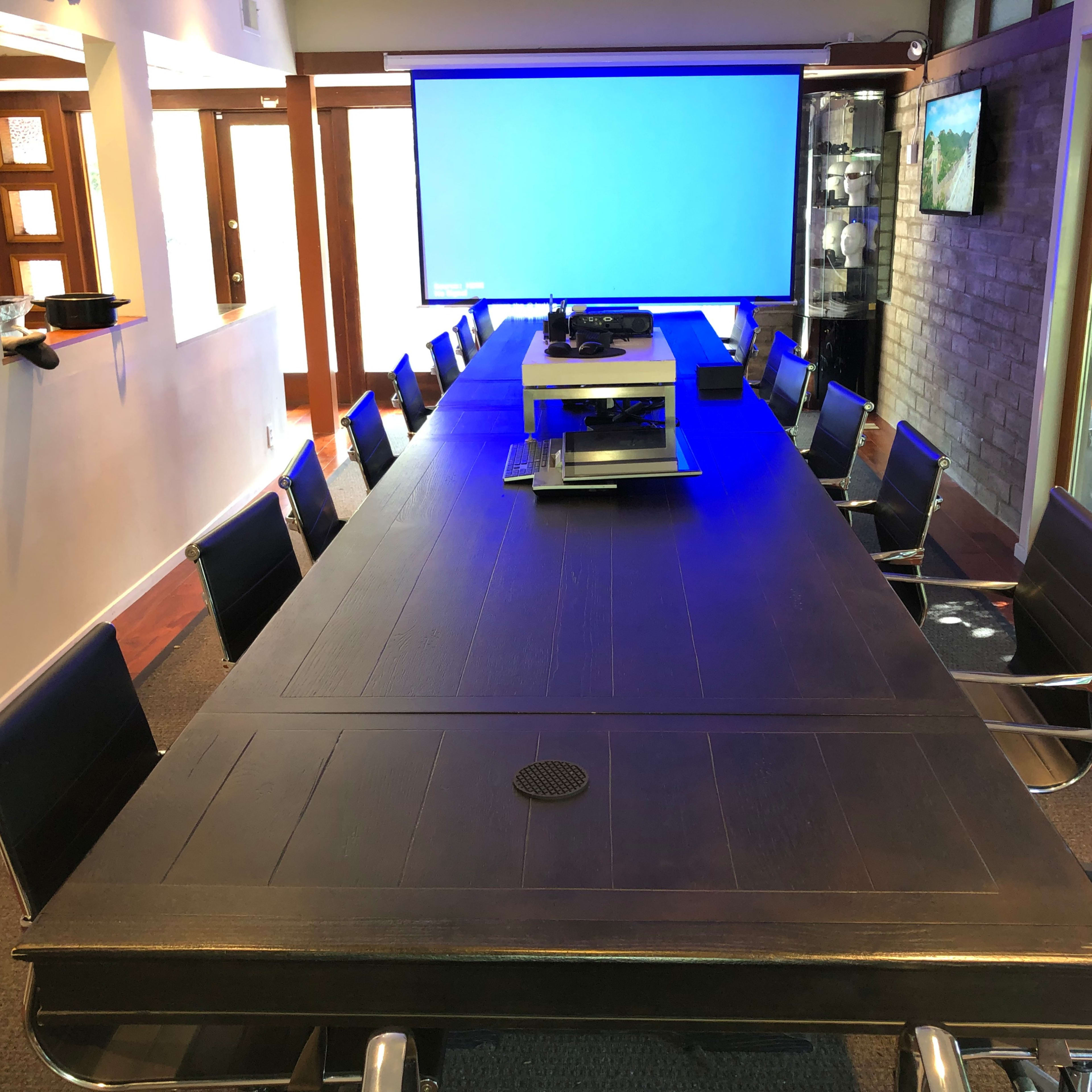 A large, long conference table is set up in a well-lit meeting room, equipped with a projector and surrounded by black chairs.