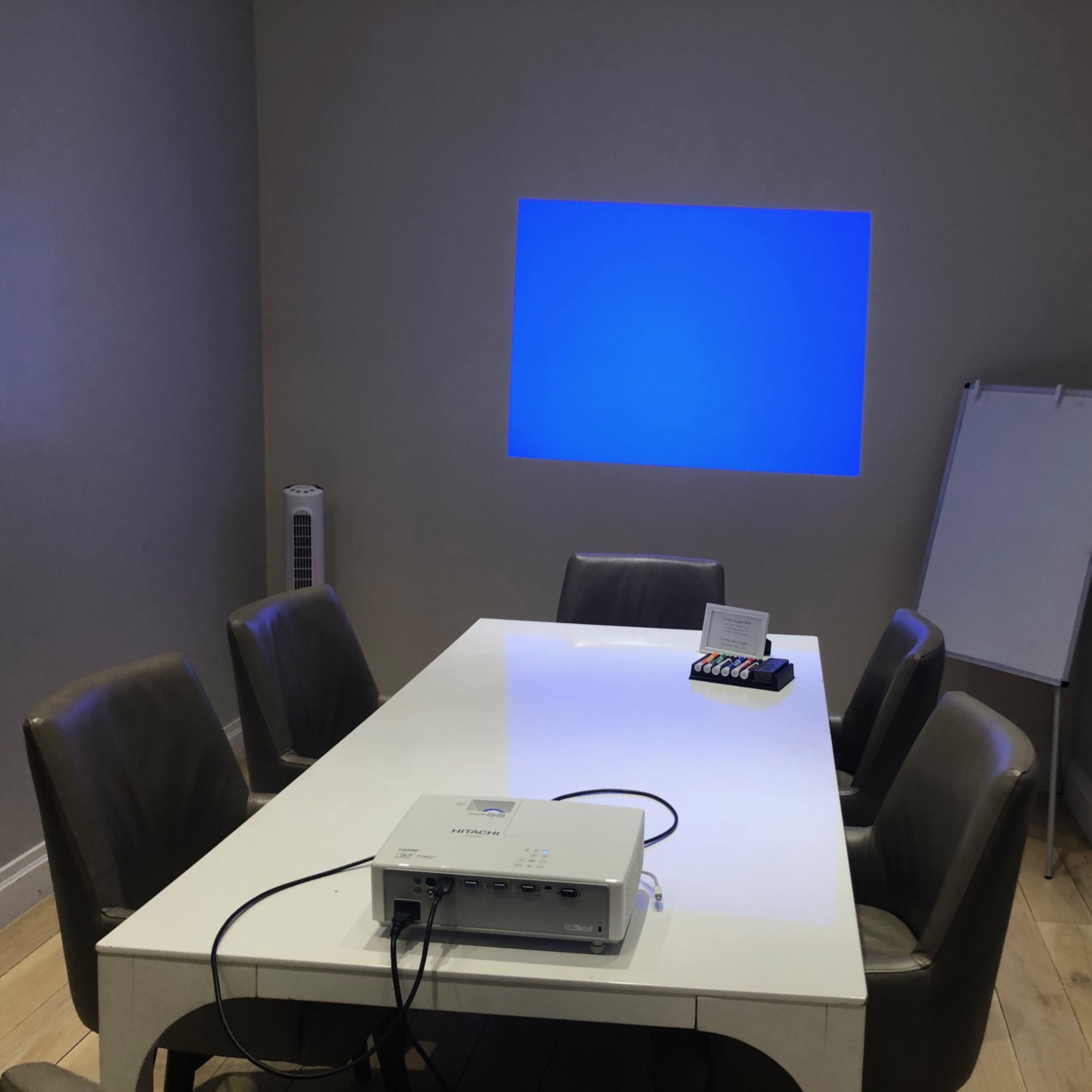 A conference room with a white table, several chairs, a projector on the table, and a blue projection on the wall.