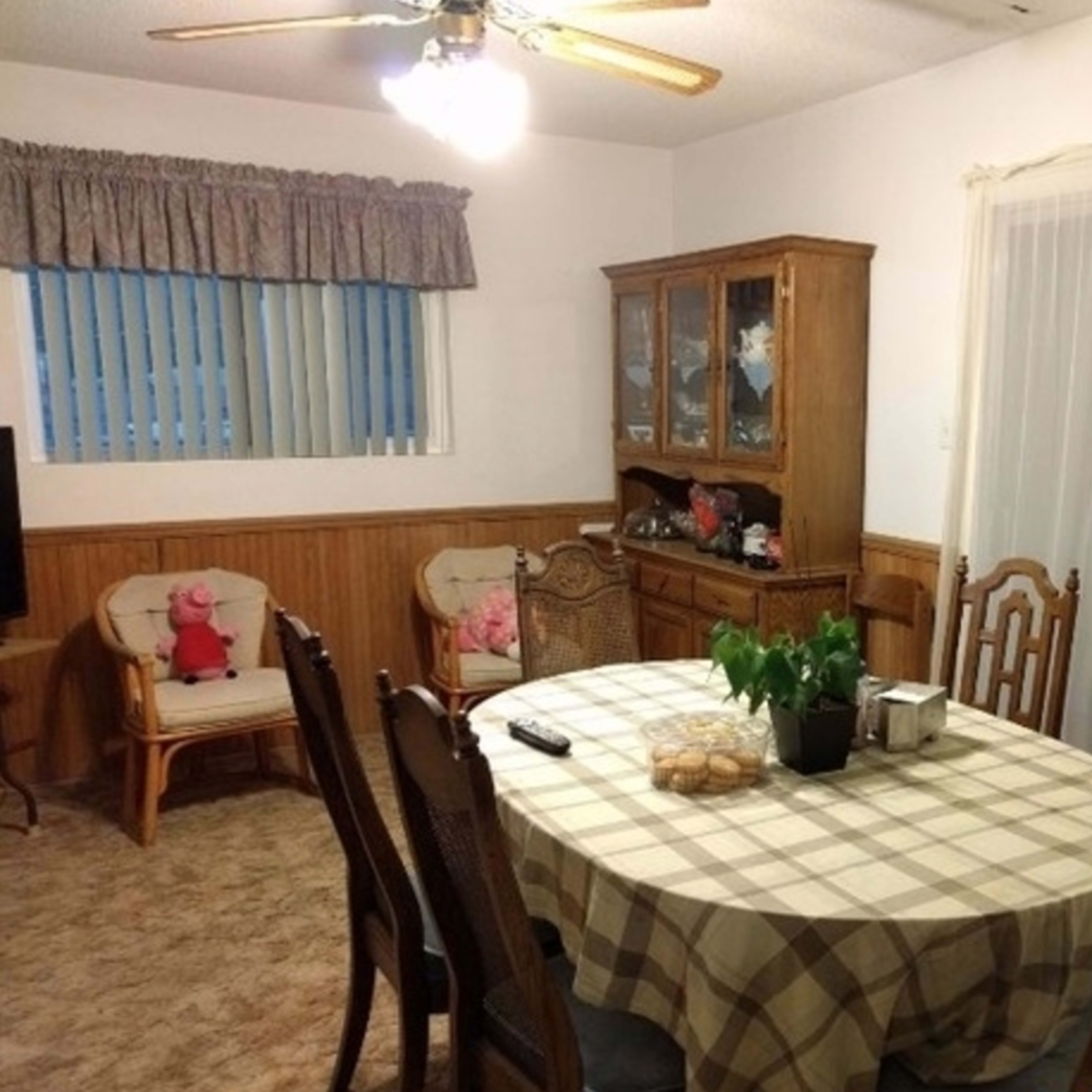 A dining area with a round table covered by a checkered tablecloth, surrounded by wooden chairs, and a china cabinet in the corner.