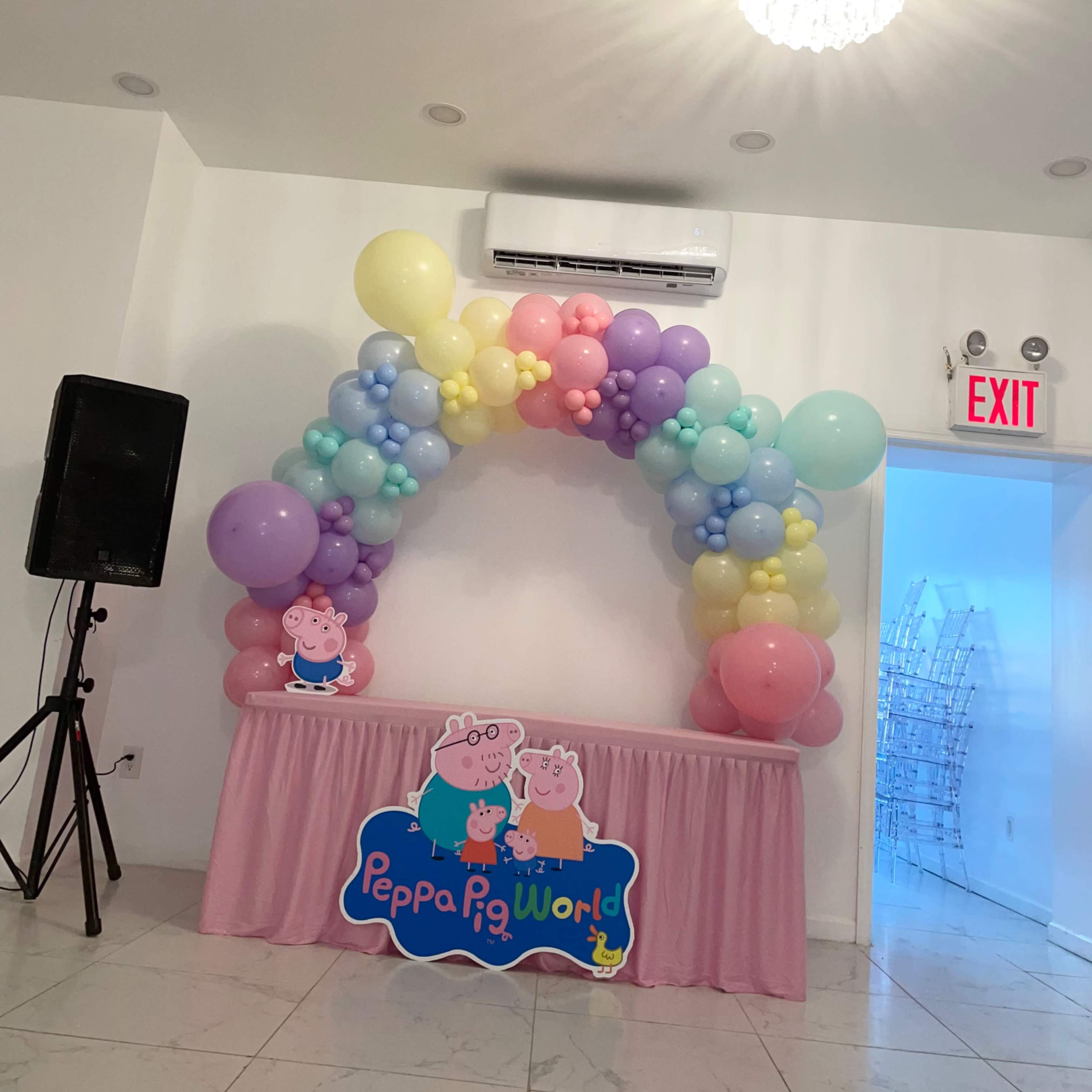 A colorful balloon arch with a pink table adorned with a "Peppa Pig World" sign is set up in a room with white walls and a chandelier.