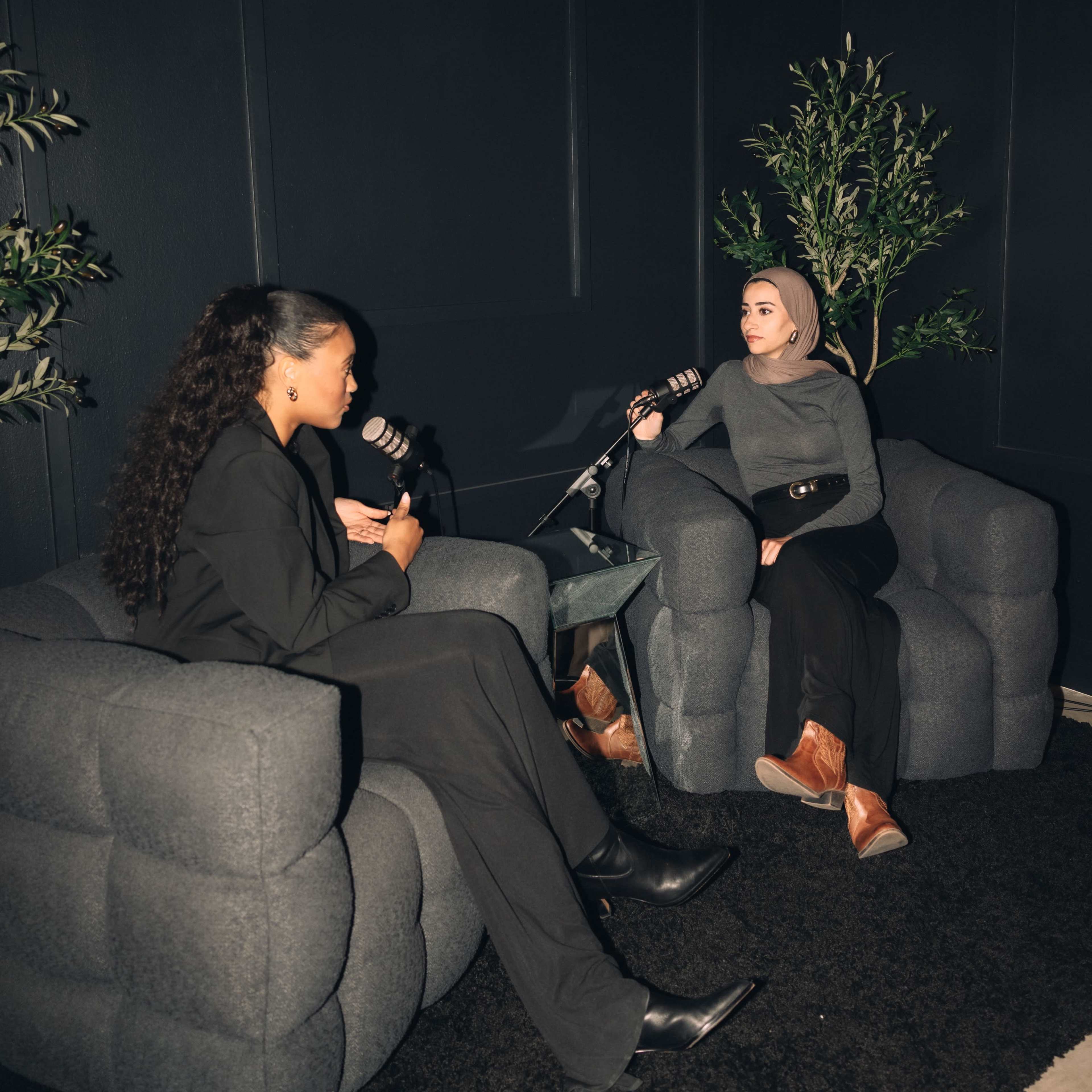 Two women sit in gray armchairs in a dark room, each holding a microphone and engaged in conversation.