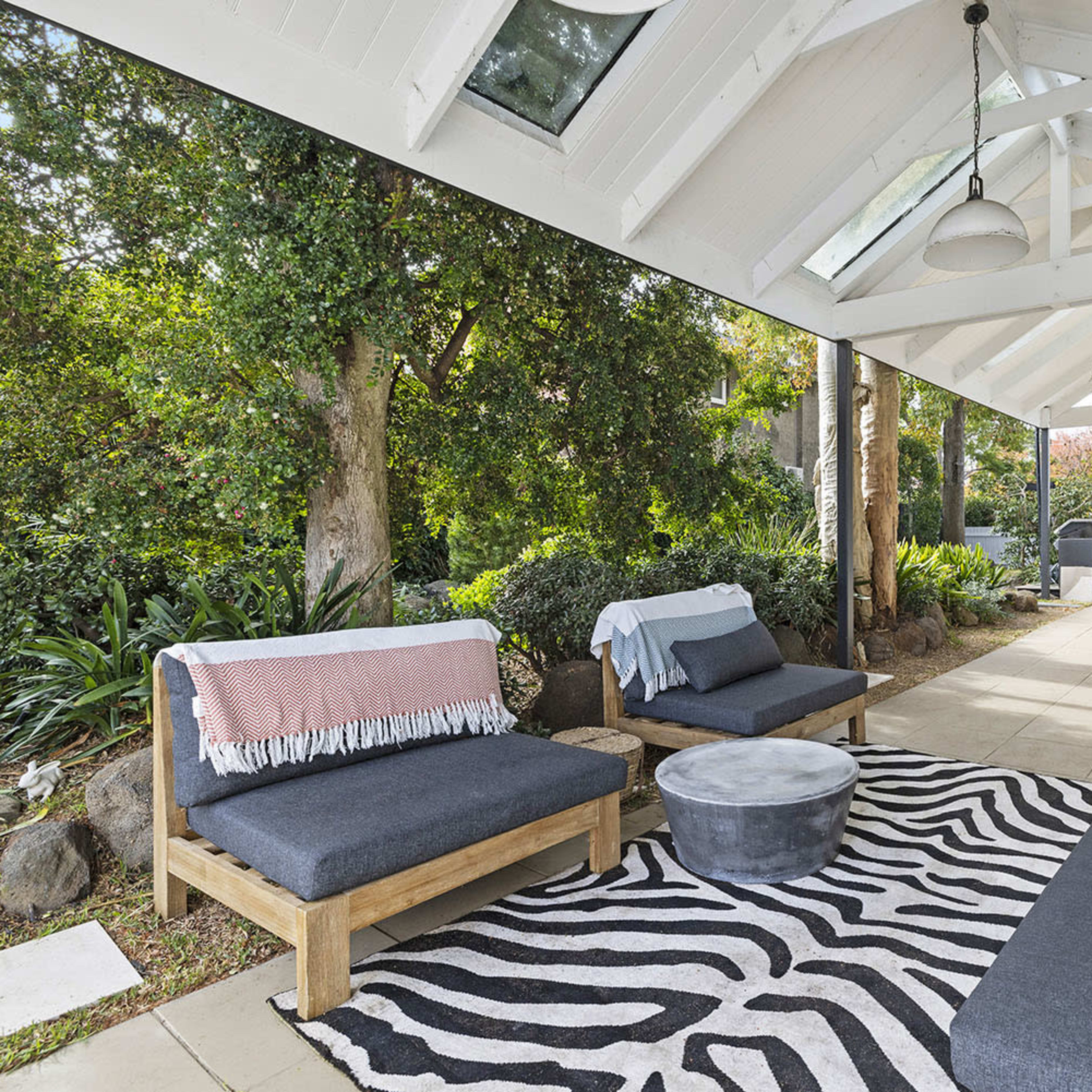 A modern outdoor living space features a seating area with wooden couches, a round coffee table, and a striped rug under a vaulted ceiling.