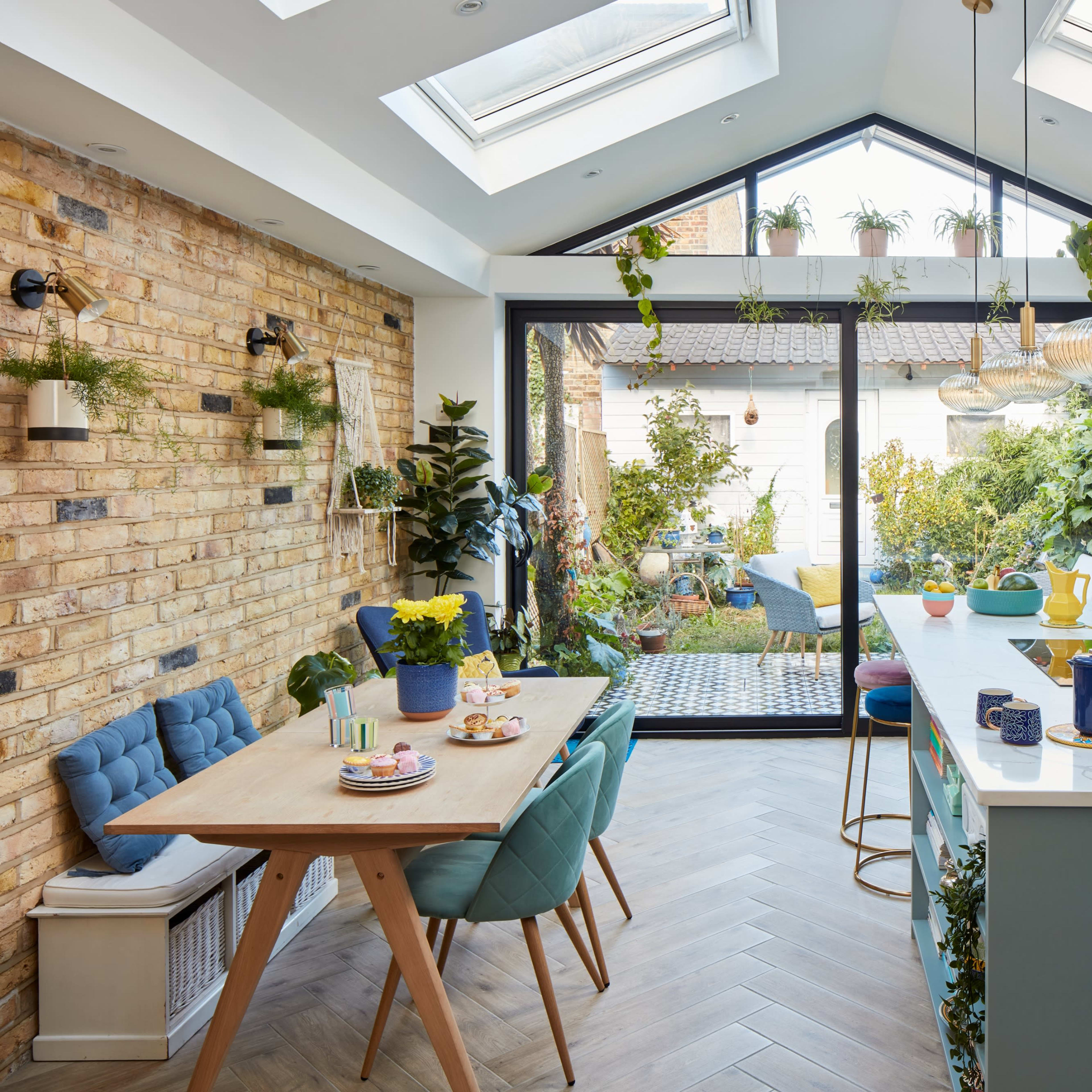 A modern kitchen with a large island, blue cabinetry, a dining table set for a meal, and abundant greenery seen through large glass doors.