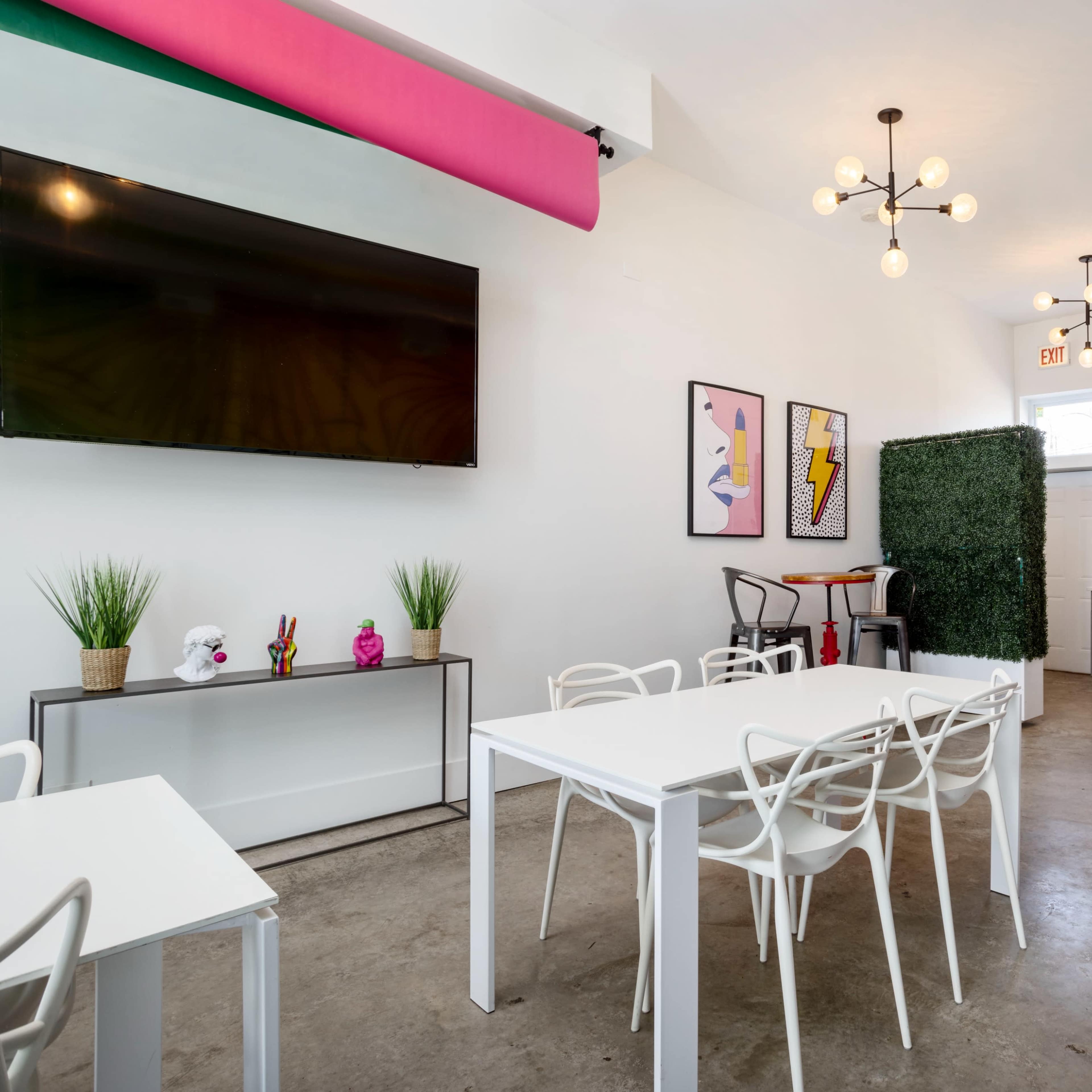 A modern, brightly lit room featuring white furniture, a large television on the wall, and a kitchenette area in the background.