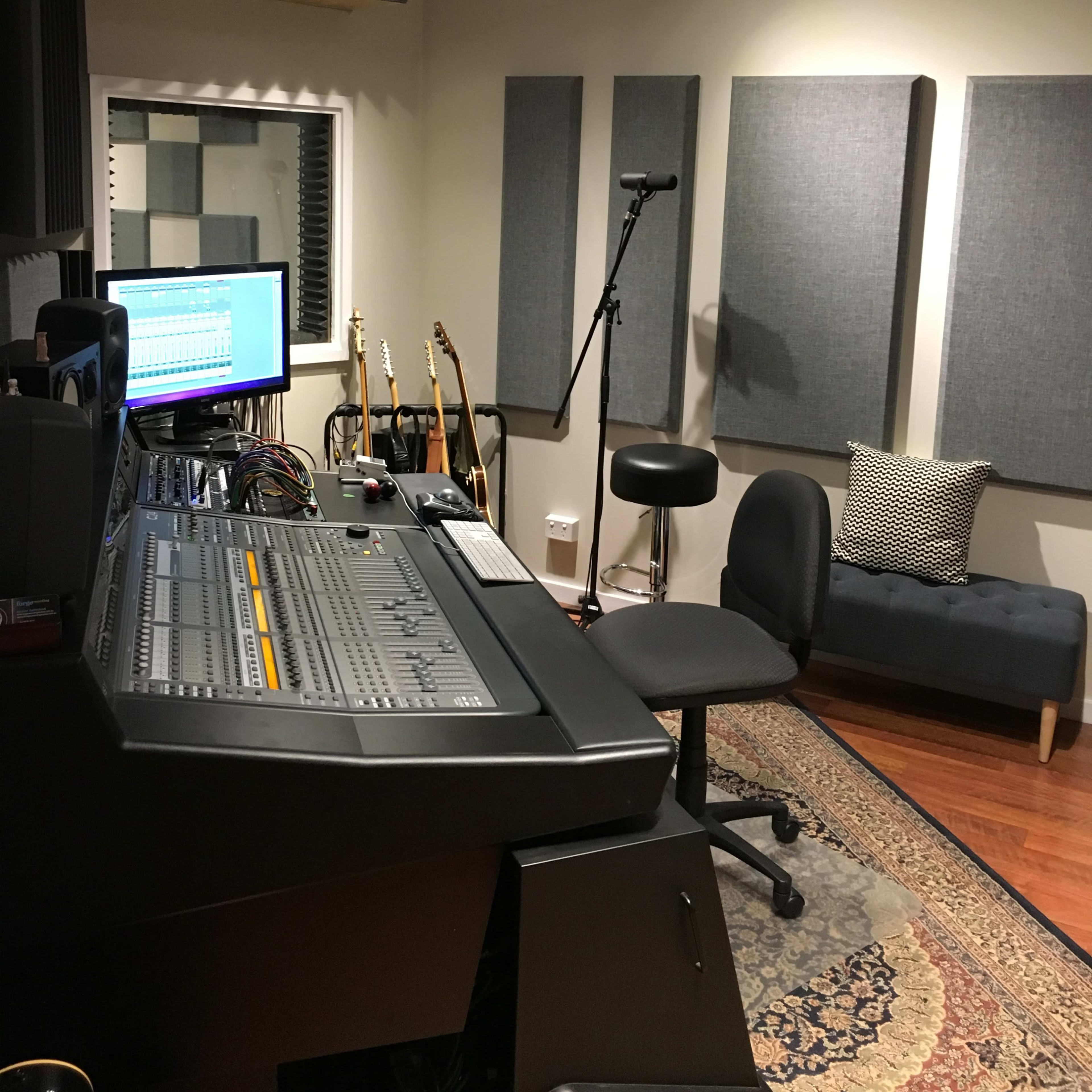 A music recording studio with a mixing console, guitars on stands, acoustic panels, and a seating area.