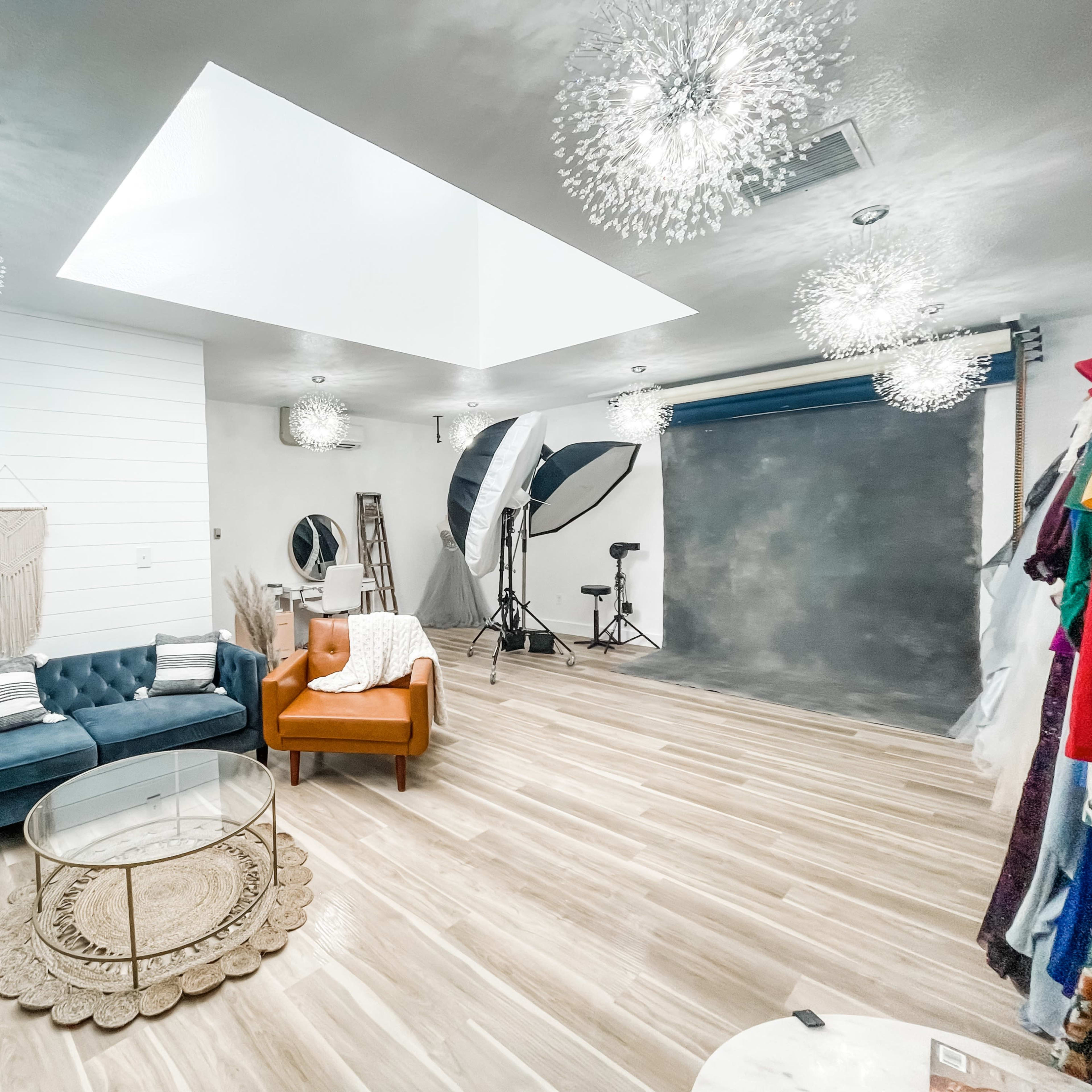 A spacious photography studio featuring a blue sofa, an orange armchair, a circular glass coffee table, and various lighting setups against a gray backdrop.