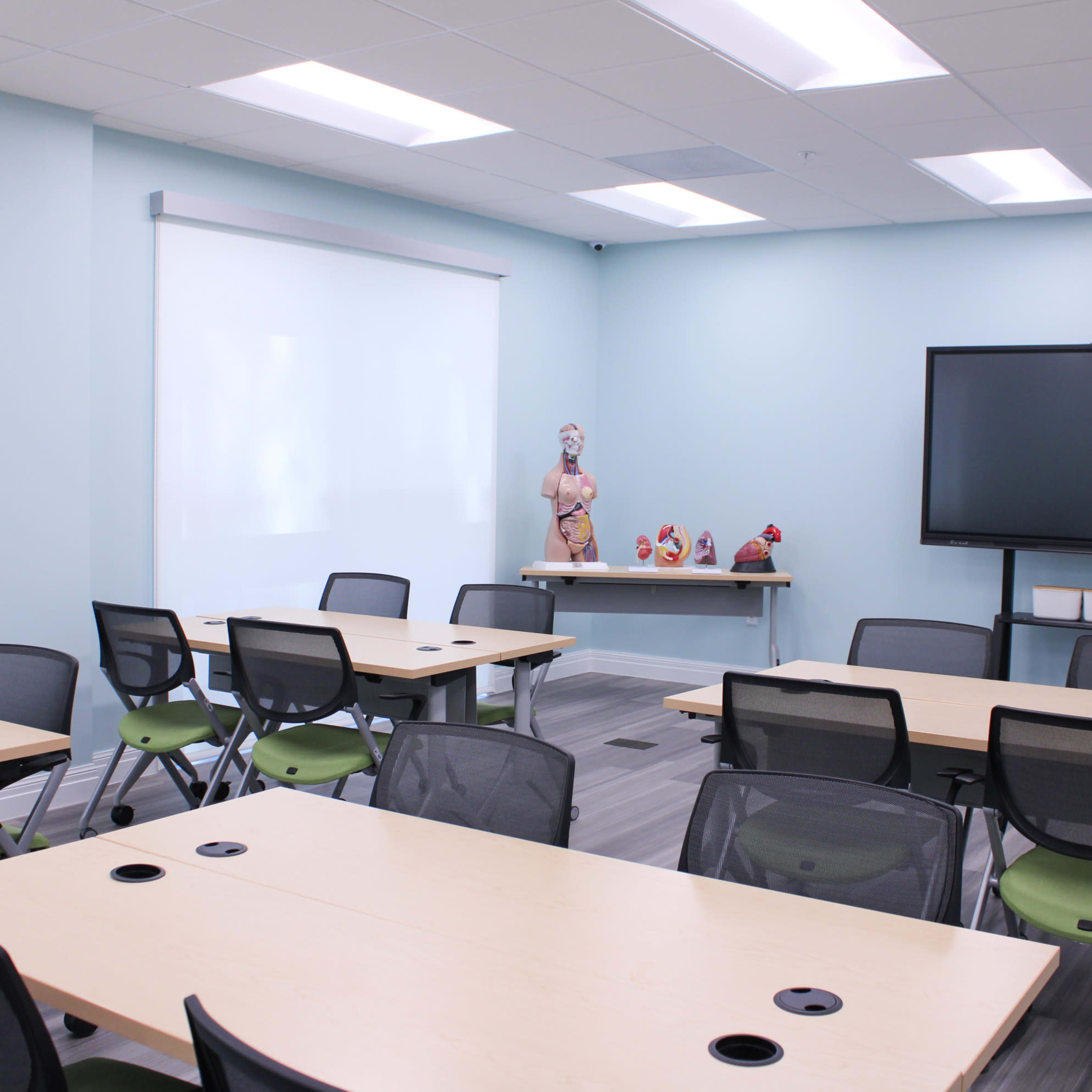 A light blue meeting room features multiple tables and chairs, a large screen, and decorative objects on a shelf.
