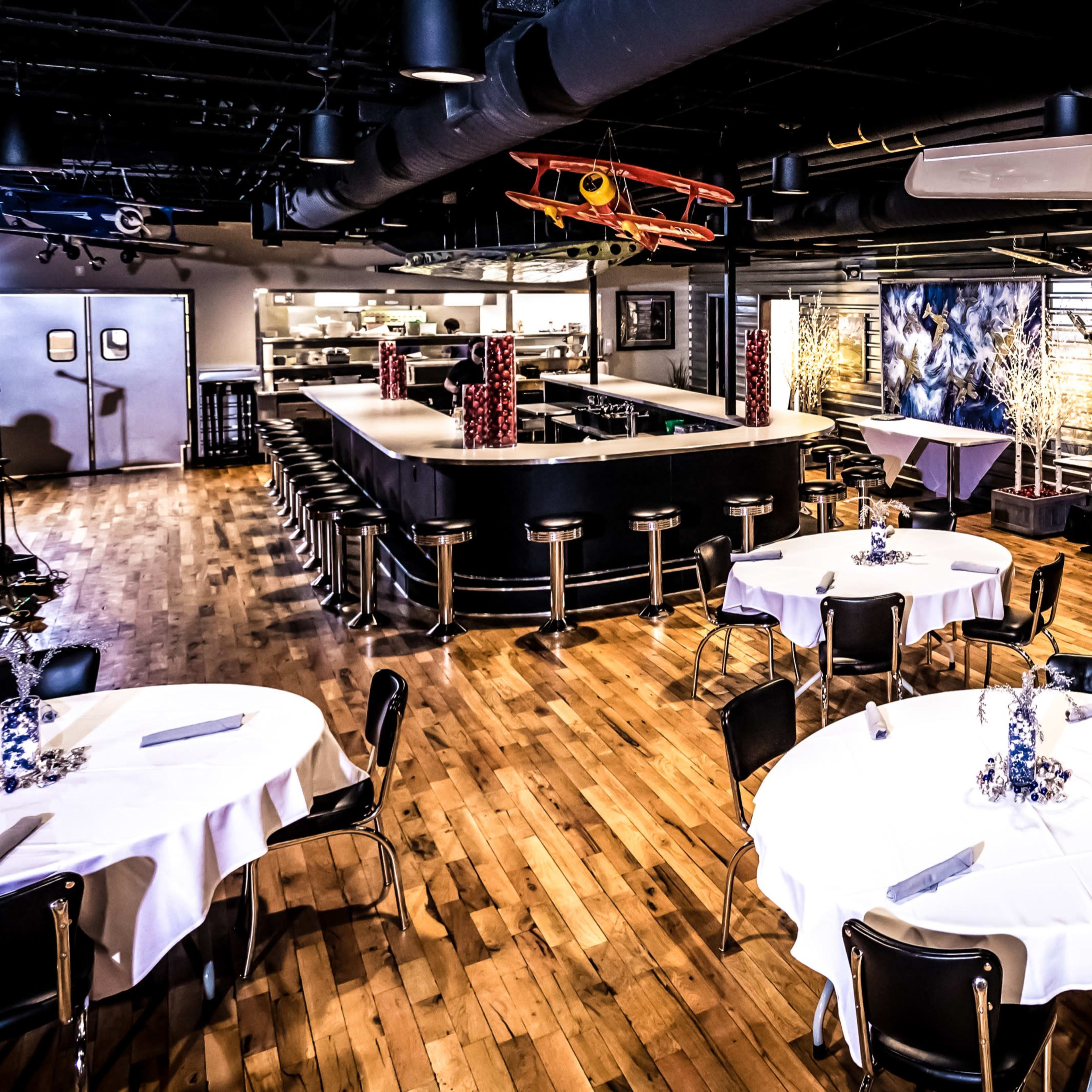 A modern restaurant interior with circular tables covered in white tablecloths, a bar area, and aviation-themed decor including a model airplane hanging from the ceiling.