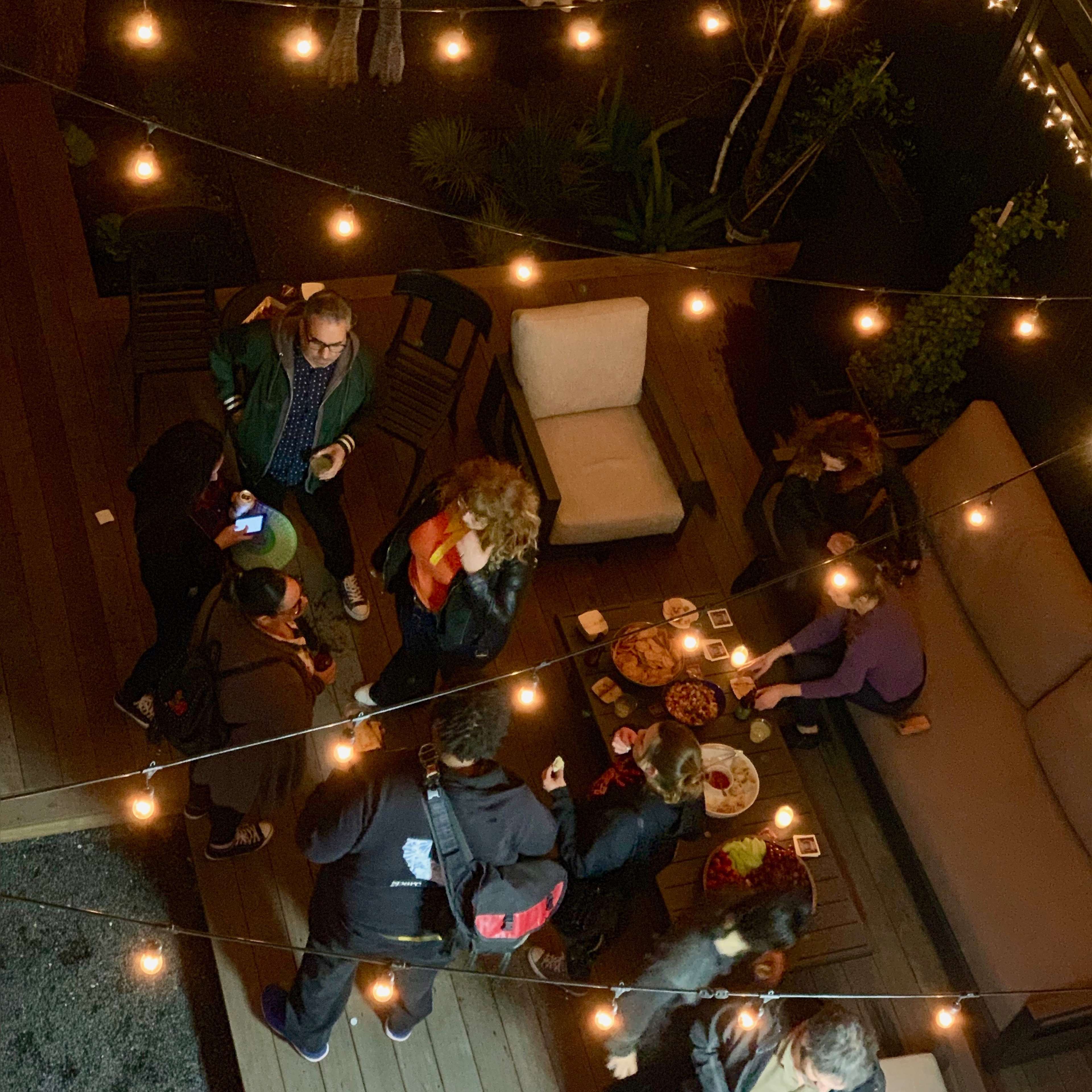A group of people gathers around tables on a wooden patio, with string lights illuminating the scene and food laid out for guests.