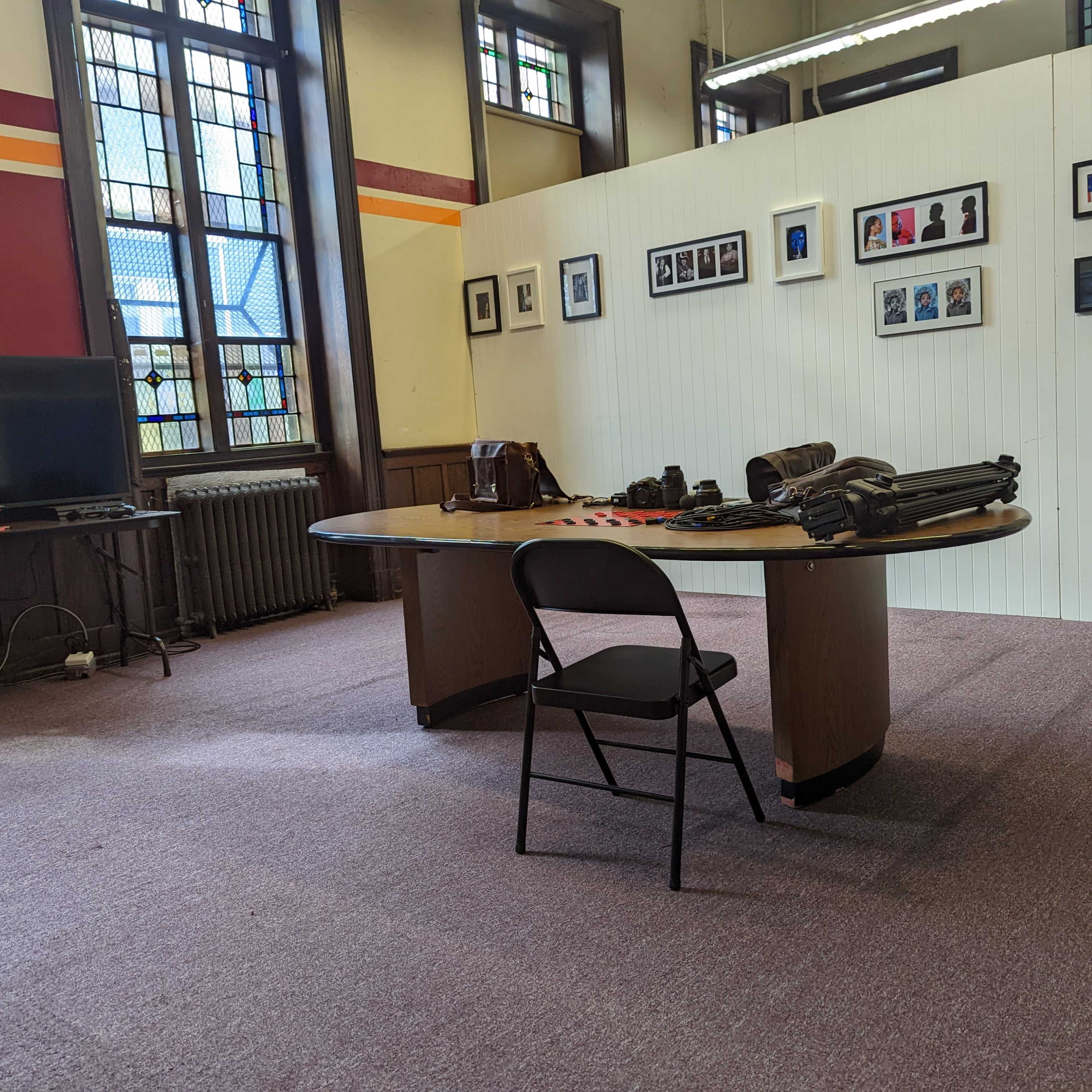 An empty room with a large table, a single chair, and several framed photos hanging on the wall.