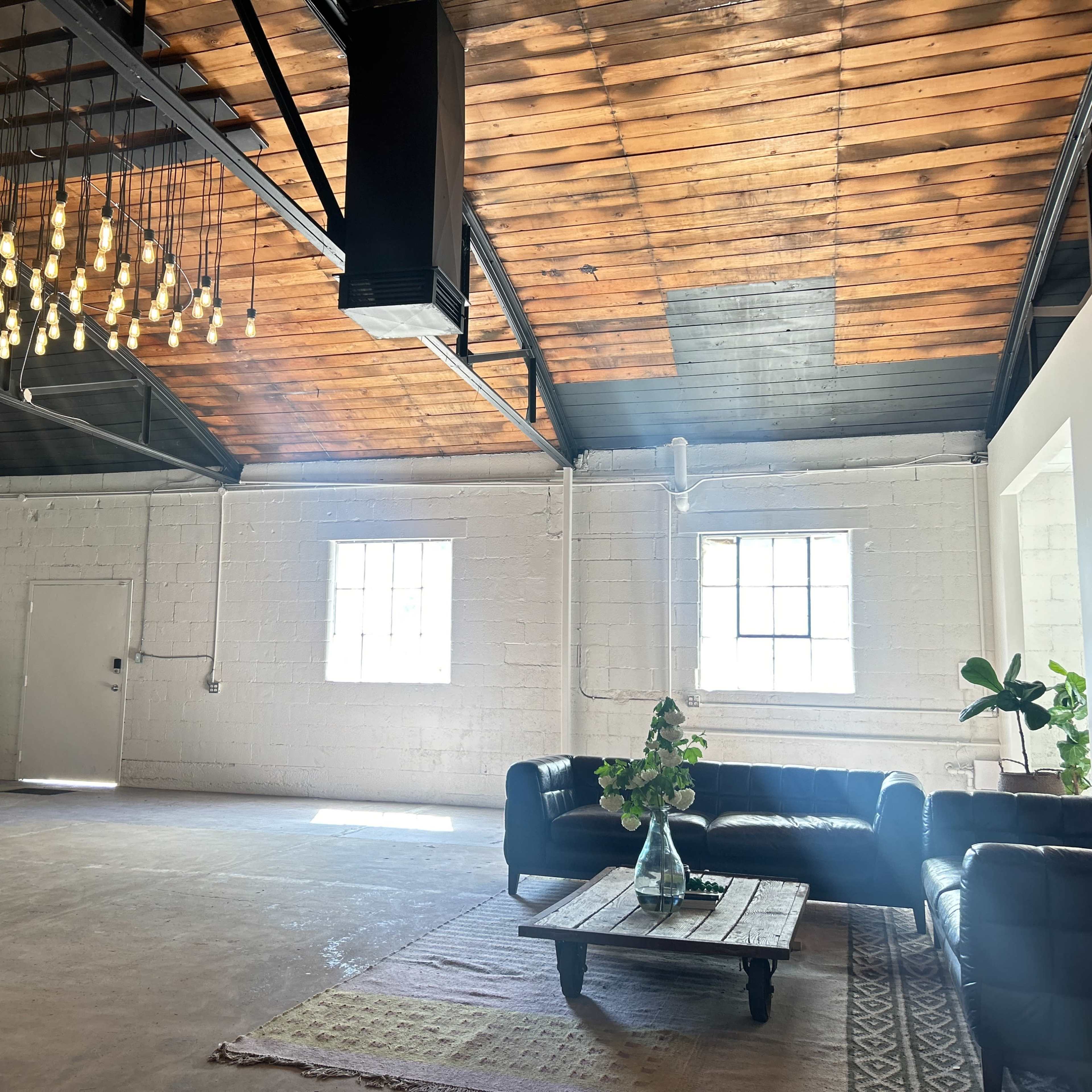 A spacious, well-lit room with a high wooden ceiling, featuring a seating area with a black sofa and a coffee table, along with potted plants and large windows.