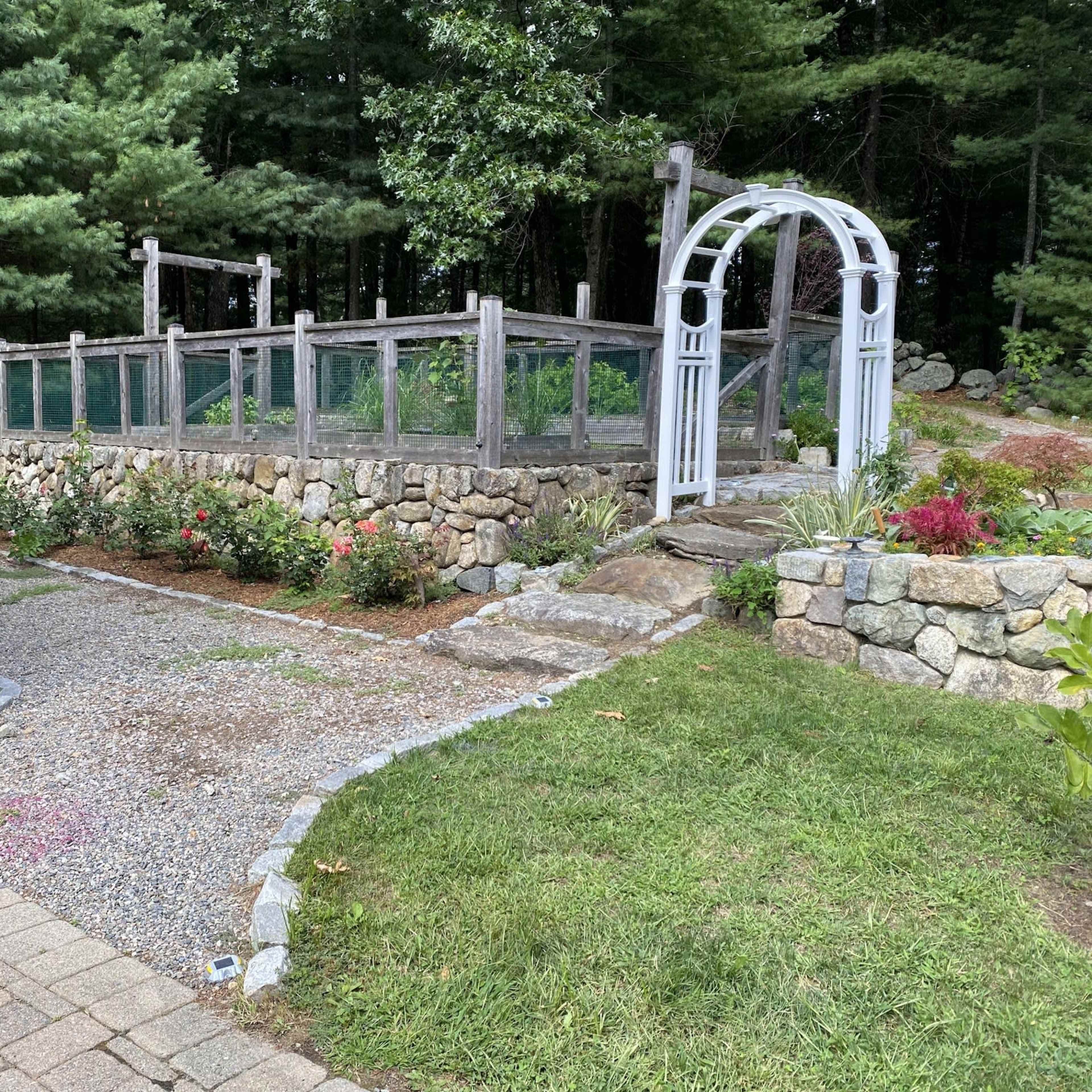 A landscaped garden area with a stone wall, wooden fencing, a white arbor, and a gravel pathway leading through the plants.
