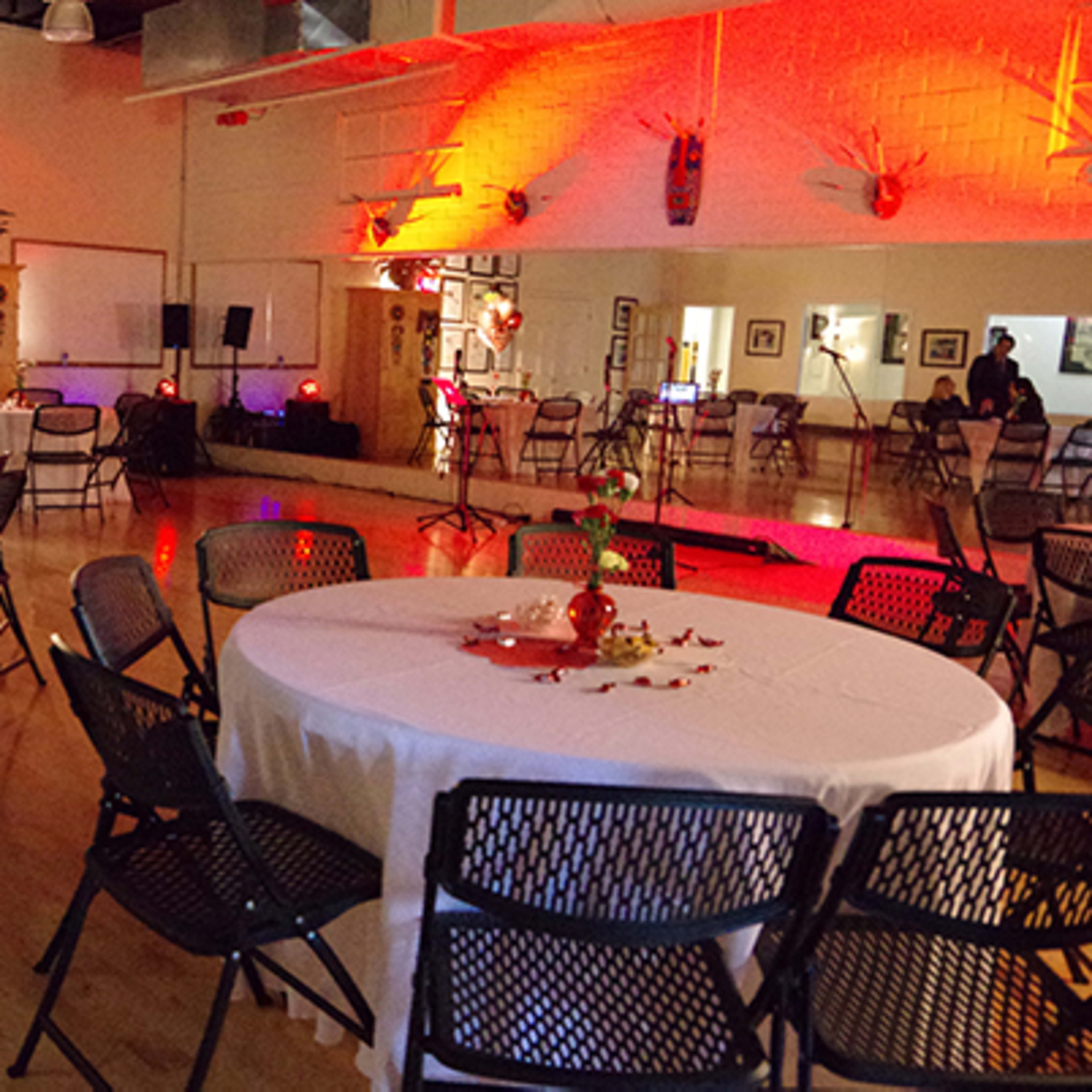 A large indoor space set up for an event, featuring round tables with white tablecloths and chairs arranged around them, with decorations and soft lighting illuminating the area.
